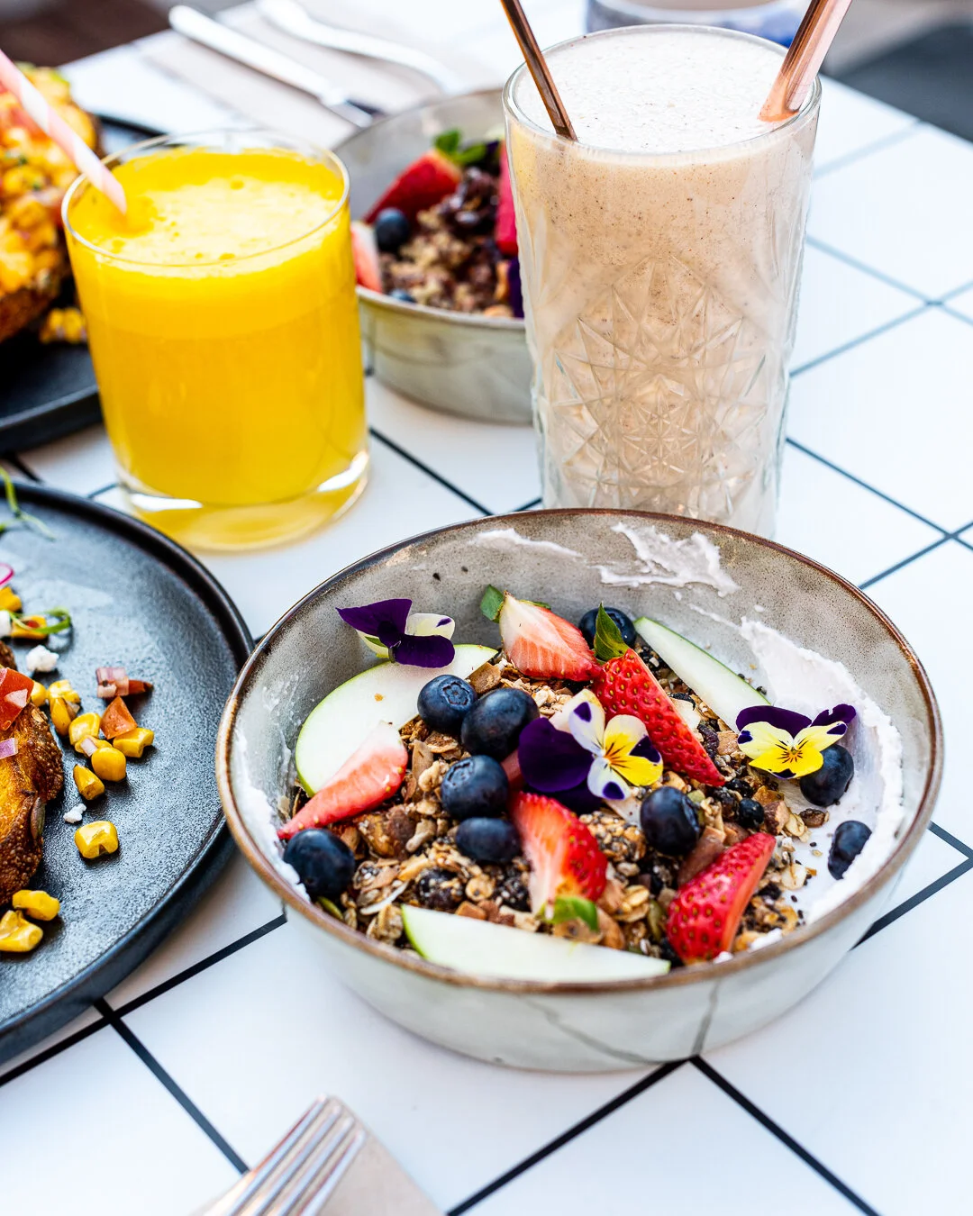A breakfast table with a bowl of granola topped with strawberries, blueberries, and edible flowers, a glass of orange juice with a straw, and a tall glass of a frothy beverage, surrounded by other breakfast dishes and a white tiled table.