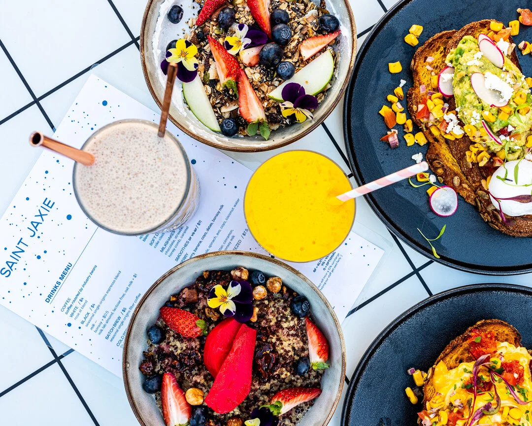 A table filled with colorful breakfast dishes including bowls of granola with berries and edible flowers, avocado toast topped with vegetables and microgreens, a smoothie, and a yellow juice, on a checkered table