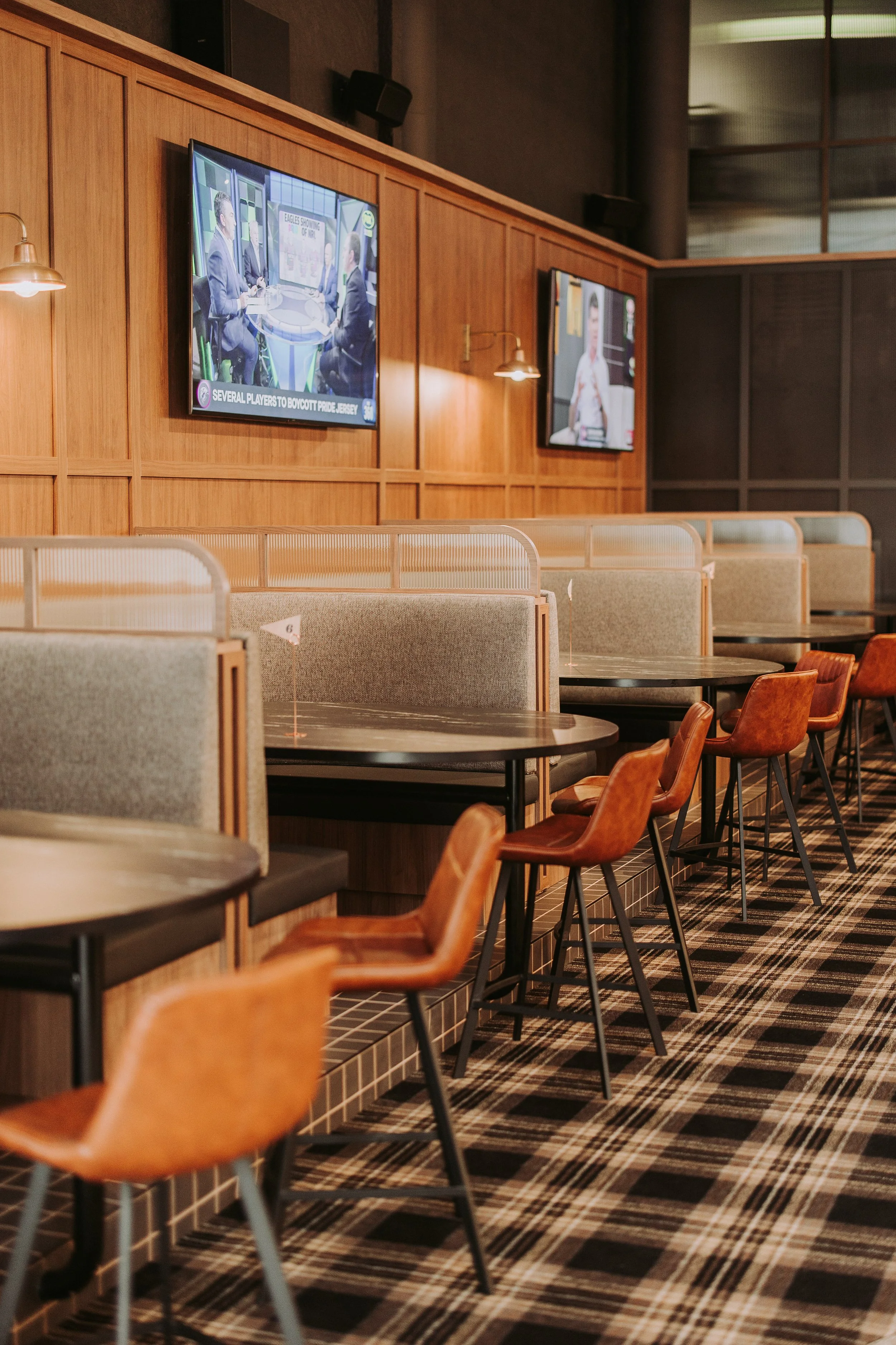 Empty seating area in a restaurant or cafe with booths and chairs, wooden panel decor, and two wall-mounted TV screens showing news or sports.
