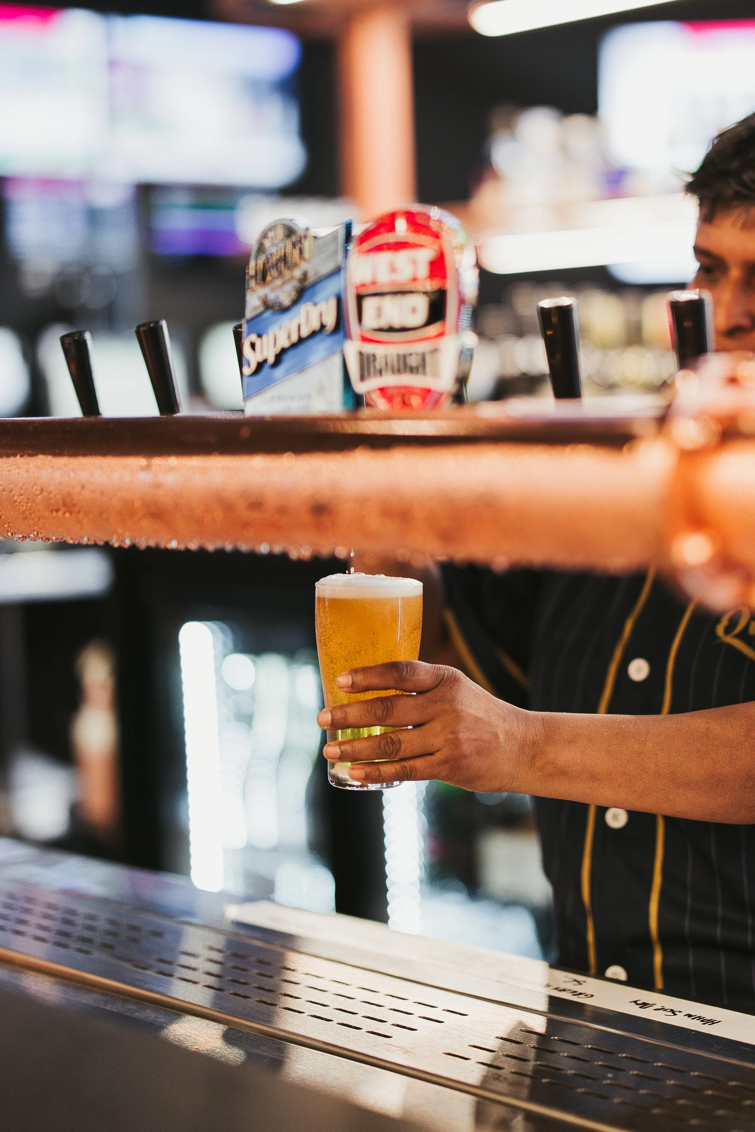 A bartender serving a glass of beer at a bar with taps and alcohol signs in the background.
