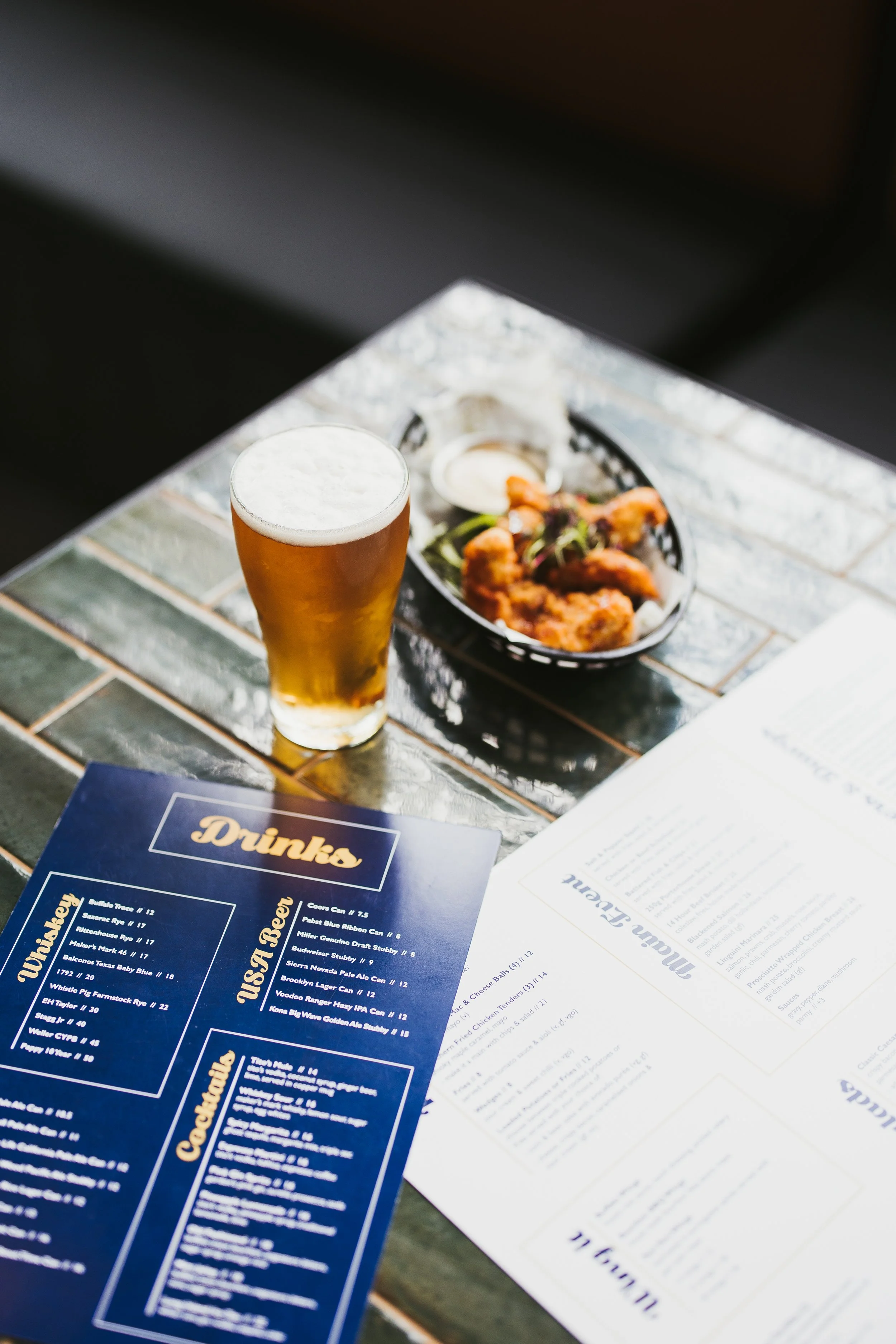 A table with a glass of beer, a basket of fried chicken wings with dipping sauce, and menus from a restaurant.