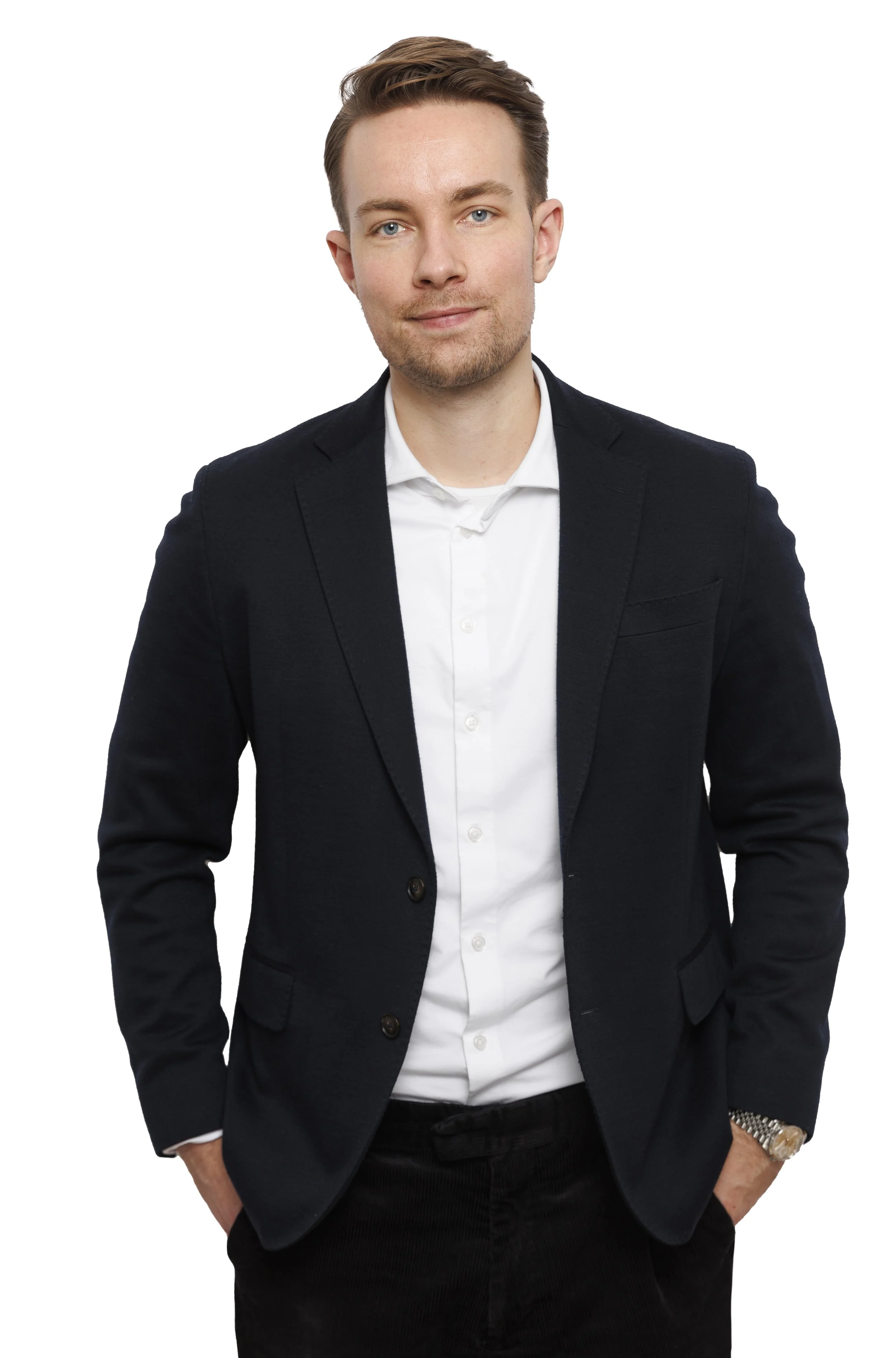 En ung mand iført sort blazer, sort t-shirt, sort bukser og ur, står med hænderne i lommerne over en hvid baggrund.