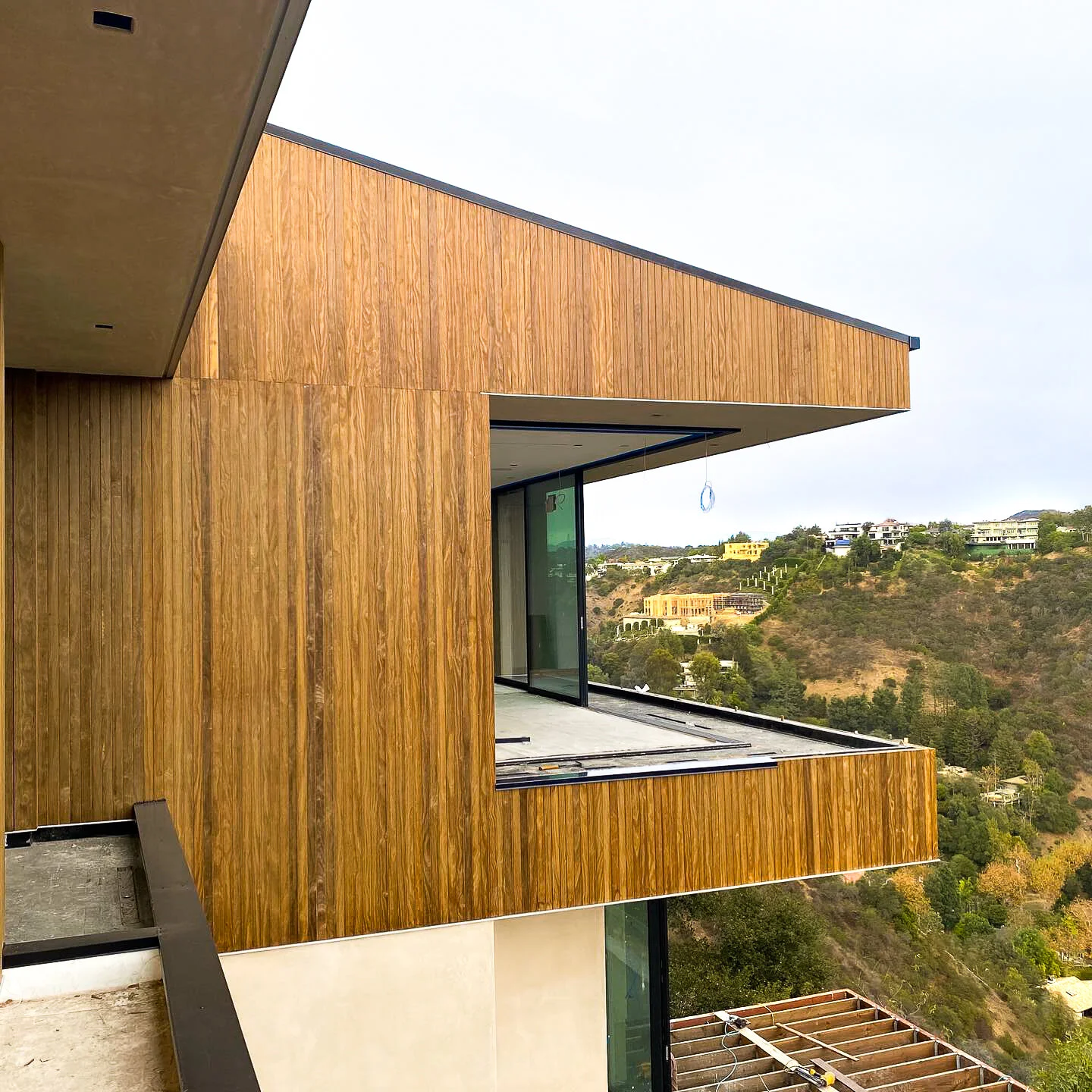 Part of a modern house under construction with wooden exterior panels and glass windows, overlooking a hilly landscape with other houses.