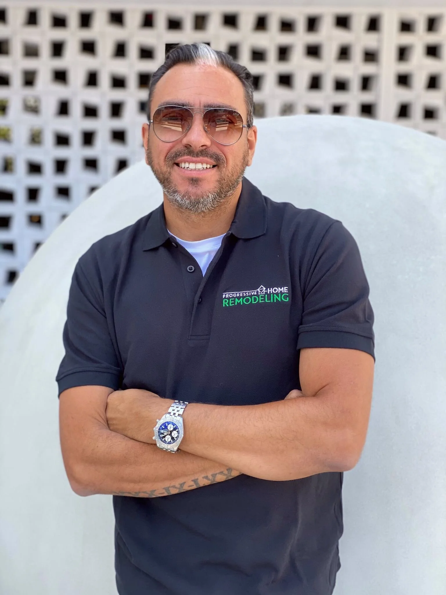 A man wearing sunglasses and a black polo shirt with a logo that says 'PROGRESSIVE HOME REMODELING', smiling with arms crossed, standing in front of a white wall with a decorative lattice pattern.