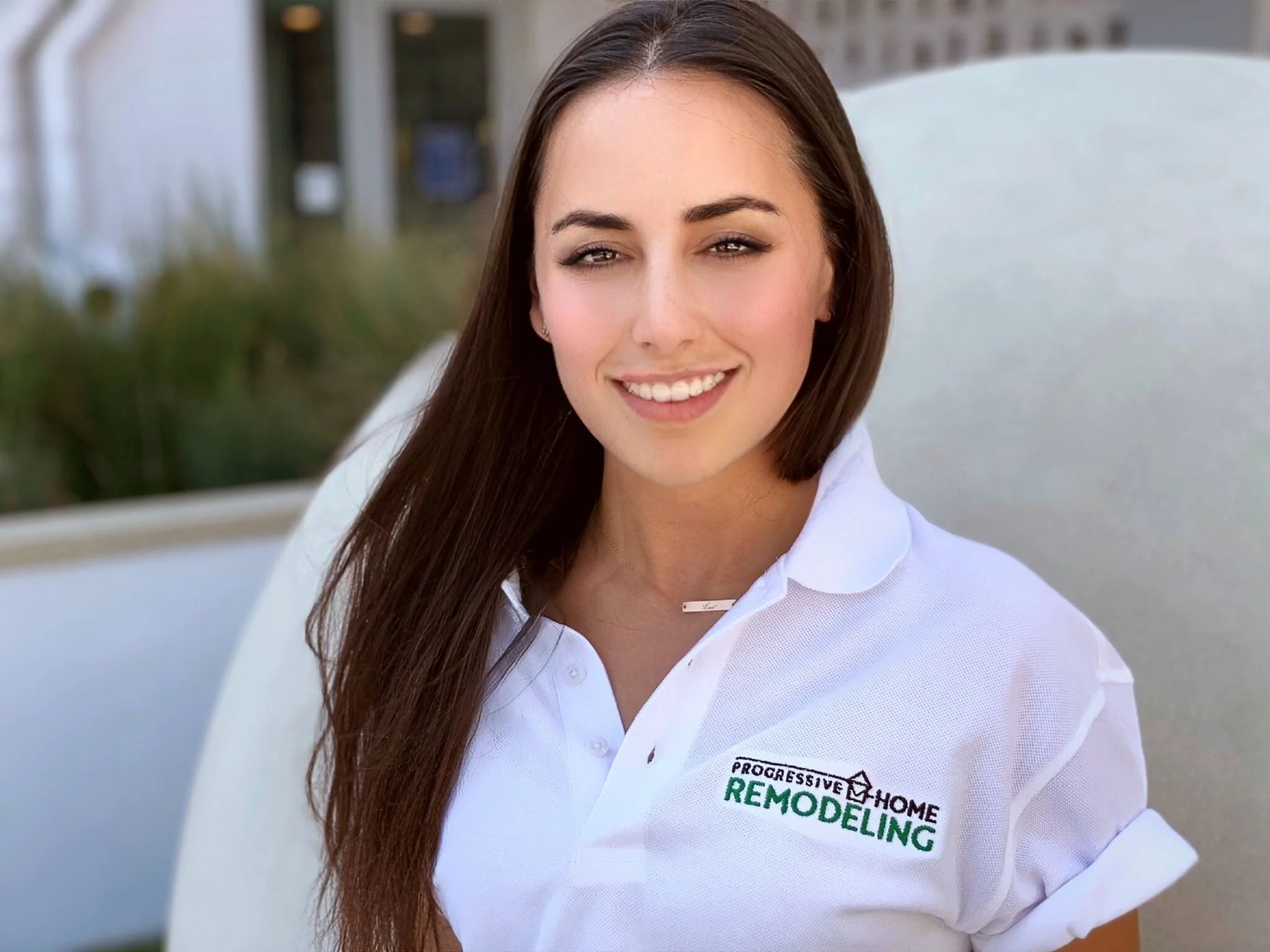 A woman with long brown hair smiling, wearing a white shirt with a logo for "Progressive Home Remodeling." She is standing outdoors, with trees and a building in the background.