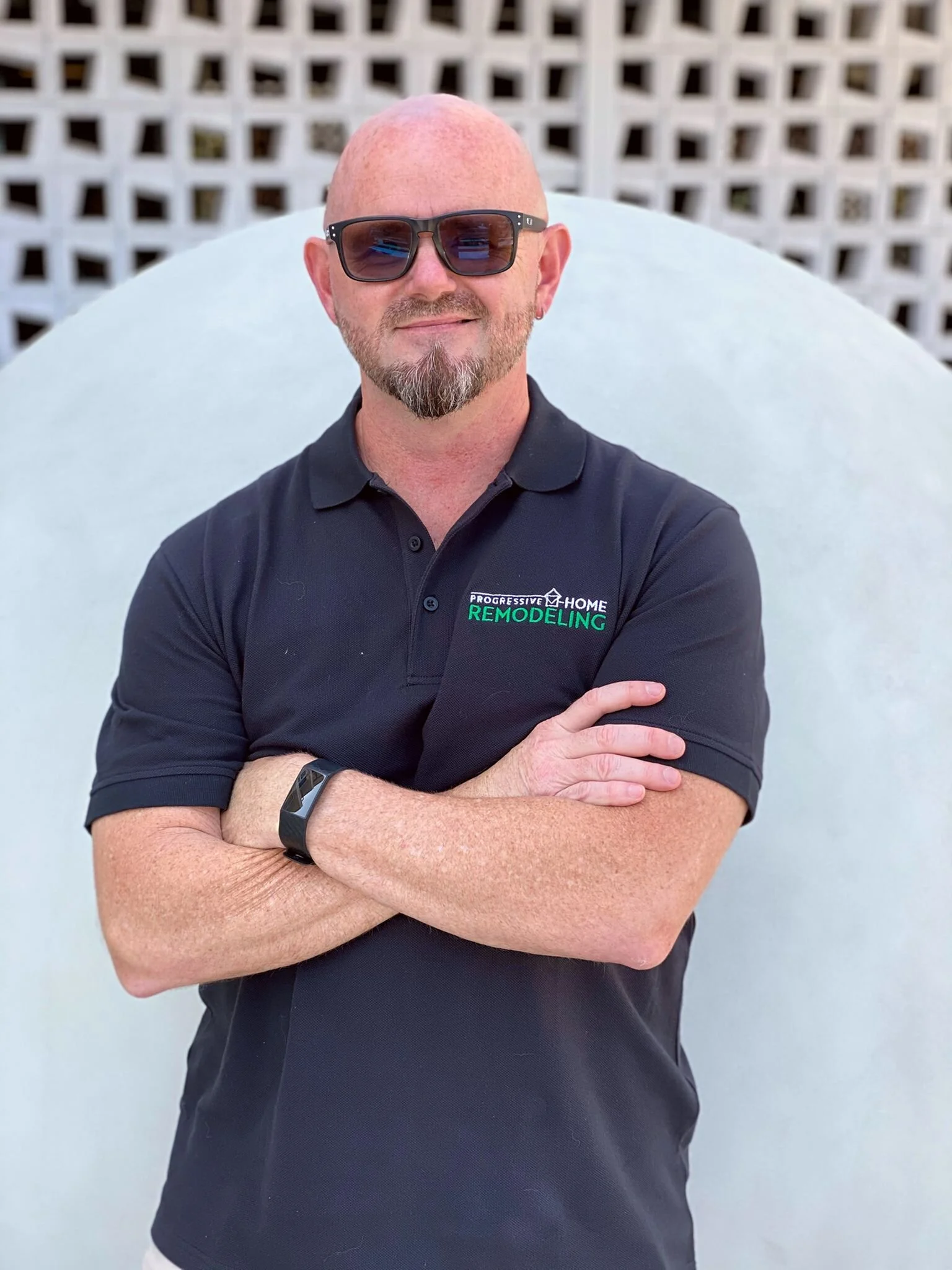 A bald man with sunglasses and a goatee, wearing a black polo shirt, standing with arms crossed in front of a white lattice background.