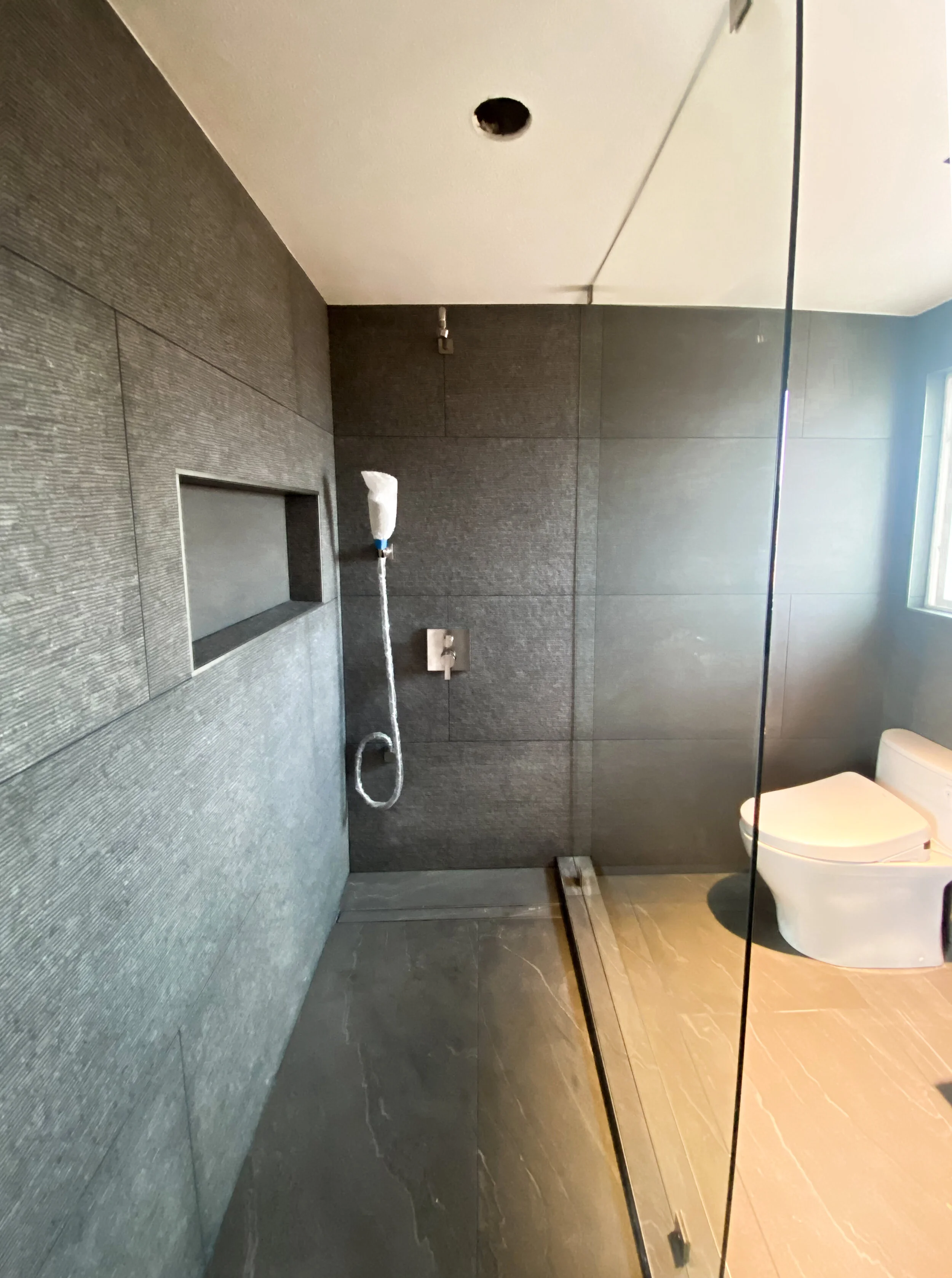 Modern bathroom with dark textured wall tiles, a white toilet, and an overhead shower with a handheld showerhead hanging from the ceiling. A narrow sink or shelf is embedded in the wall, and a window is visible on the right side.