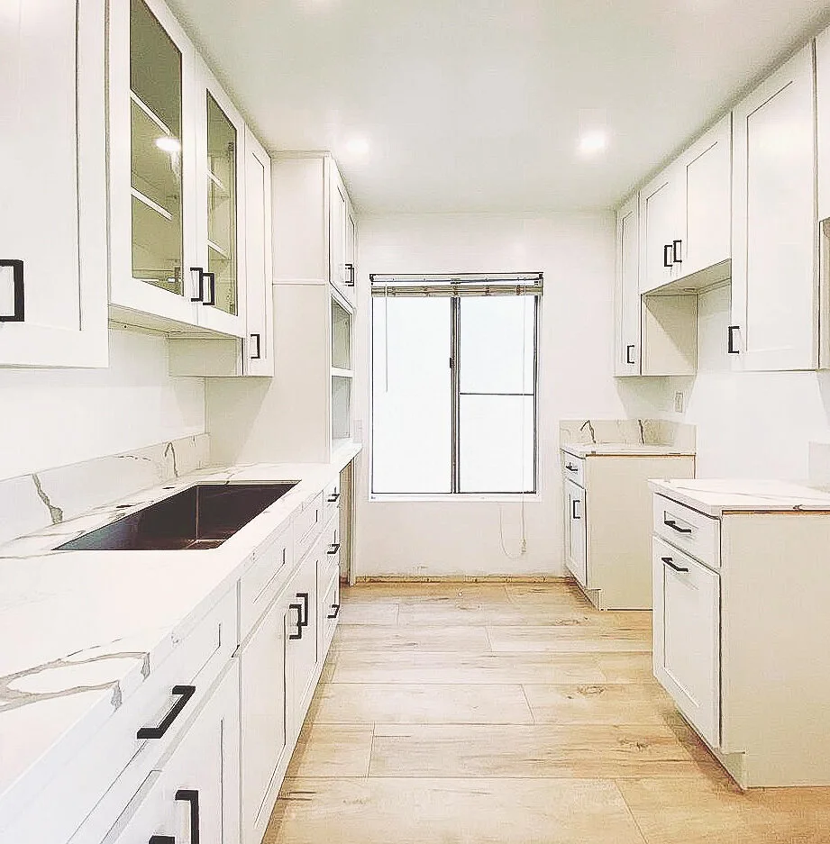 Bright white kitchen with cabinetry, black handles, a black sink, wood flooring, and a window at the back.