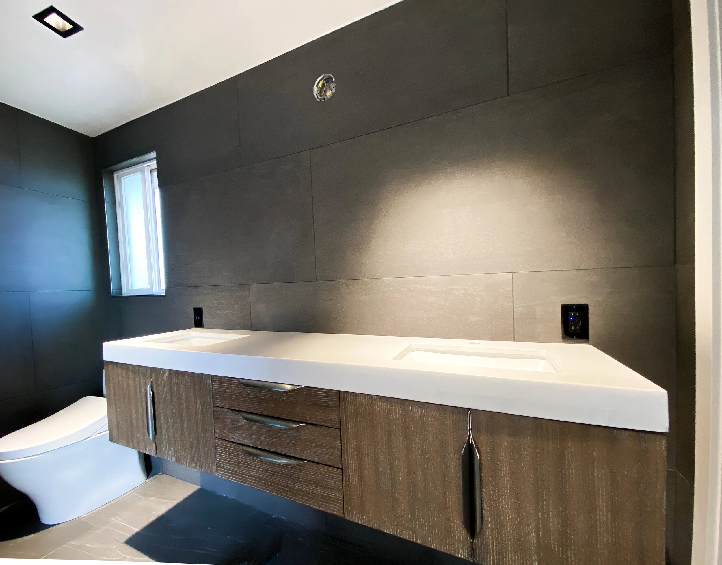 Modern bathroom with dark wooden vanity, gray tiled wall, small window, and a white bathtub.