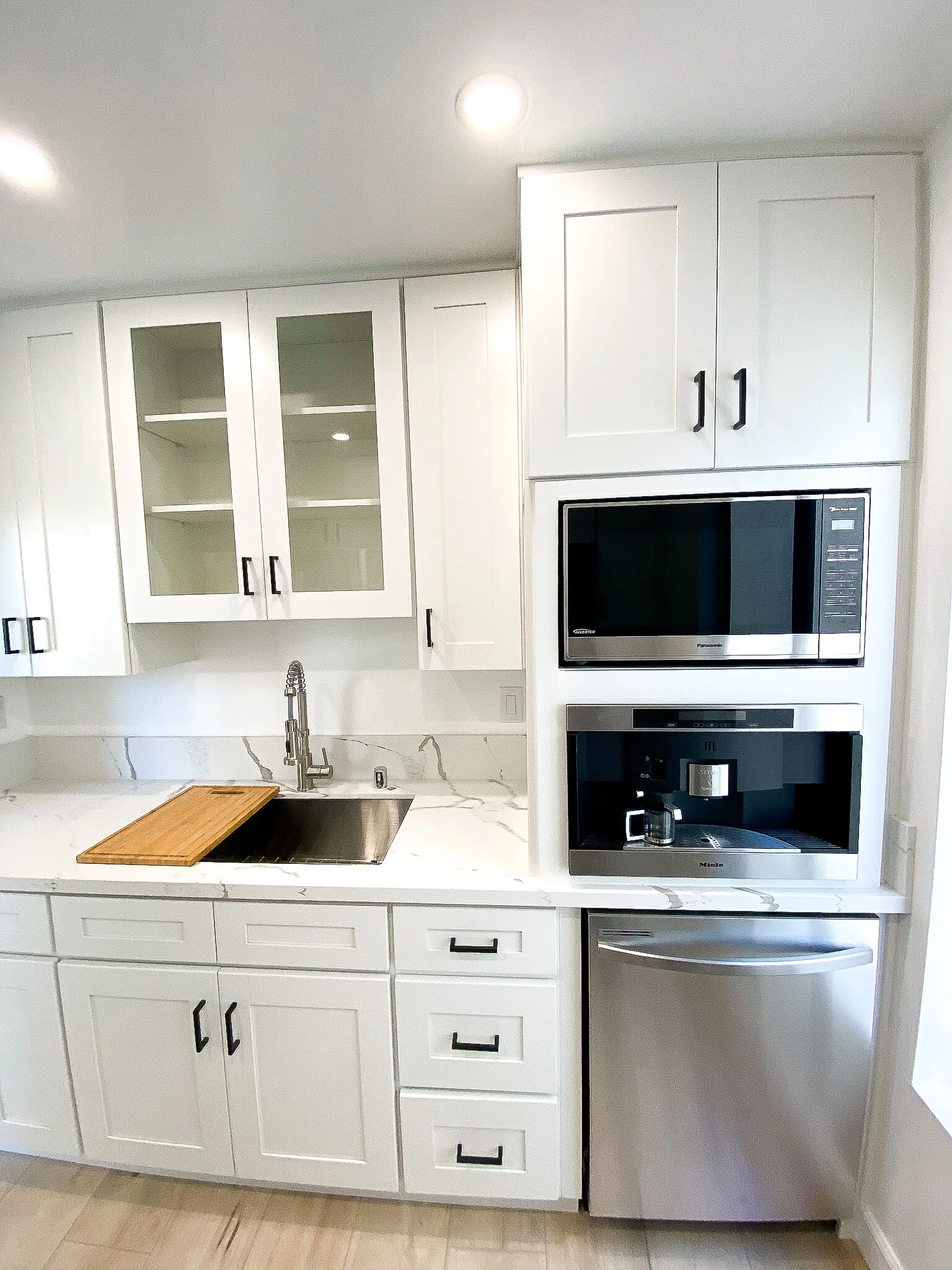 Modern white kitchen with upper cabinets, some with glass doors, lower cabinets, a marble countertop, a black microwave and oven, a coffee machine, a wooden cutting board, and a stainless steel sink.
