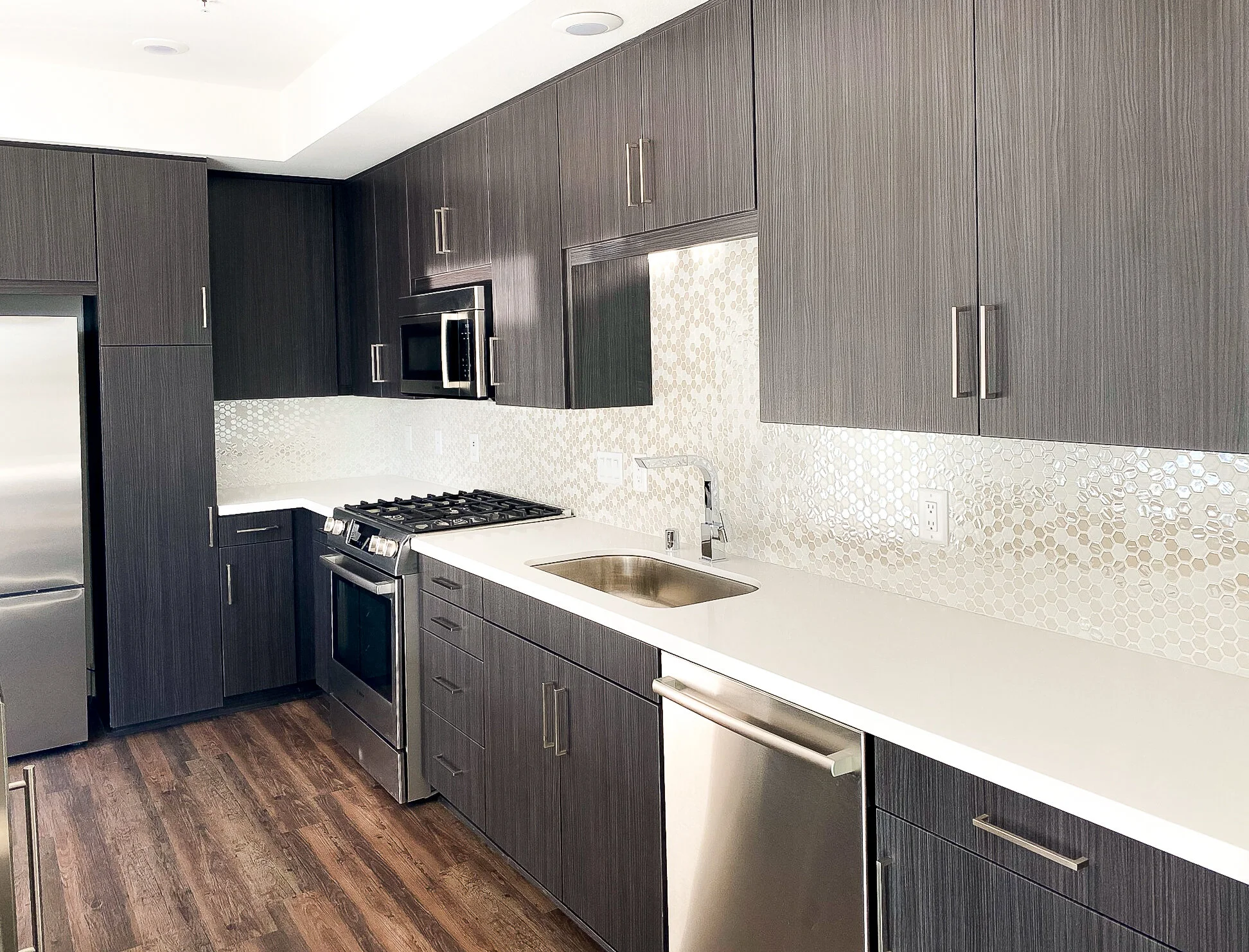 Modern kitchen with dark gray cabinets, white countertops, a stainless steel oven and microwave, and a honeycomb patterned tile backsplash, with a stainless steel refrigerator and a wood floor.