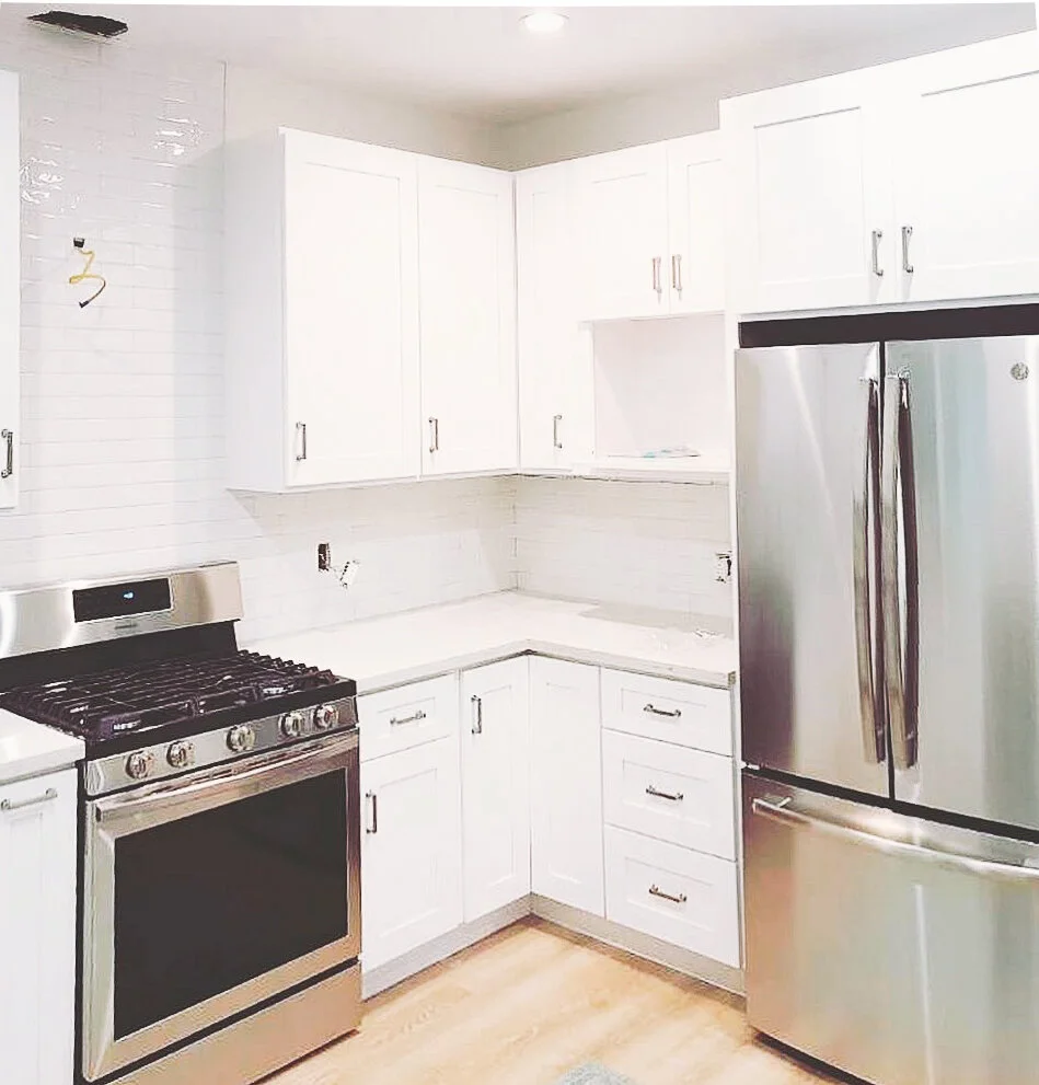 A modern kitchen with white cabinets, stainless steel appliances including a refrigerator and oven, and a light-colored hardwood floor.