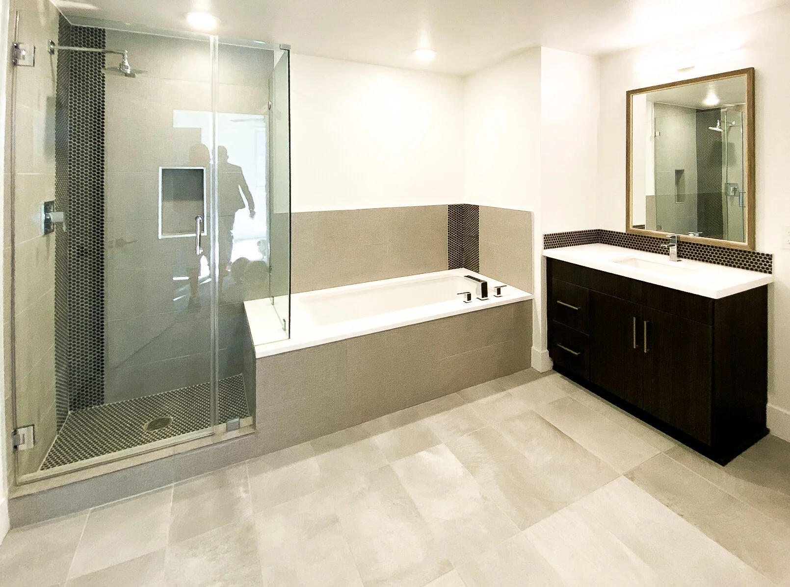 Modern bathroom with a glass-enclosed shower on the left, a built-in bathtub in the center, and a dark wood vanity with a white countertop and oval mirror on the right.