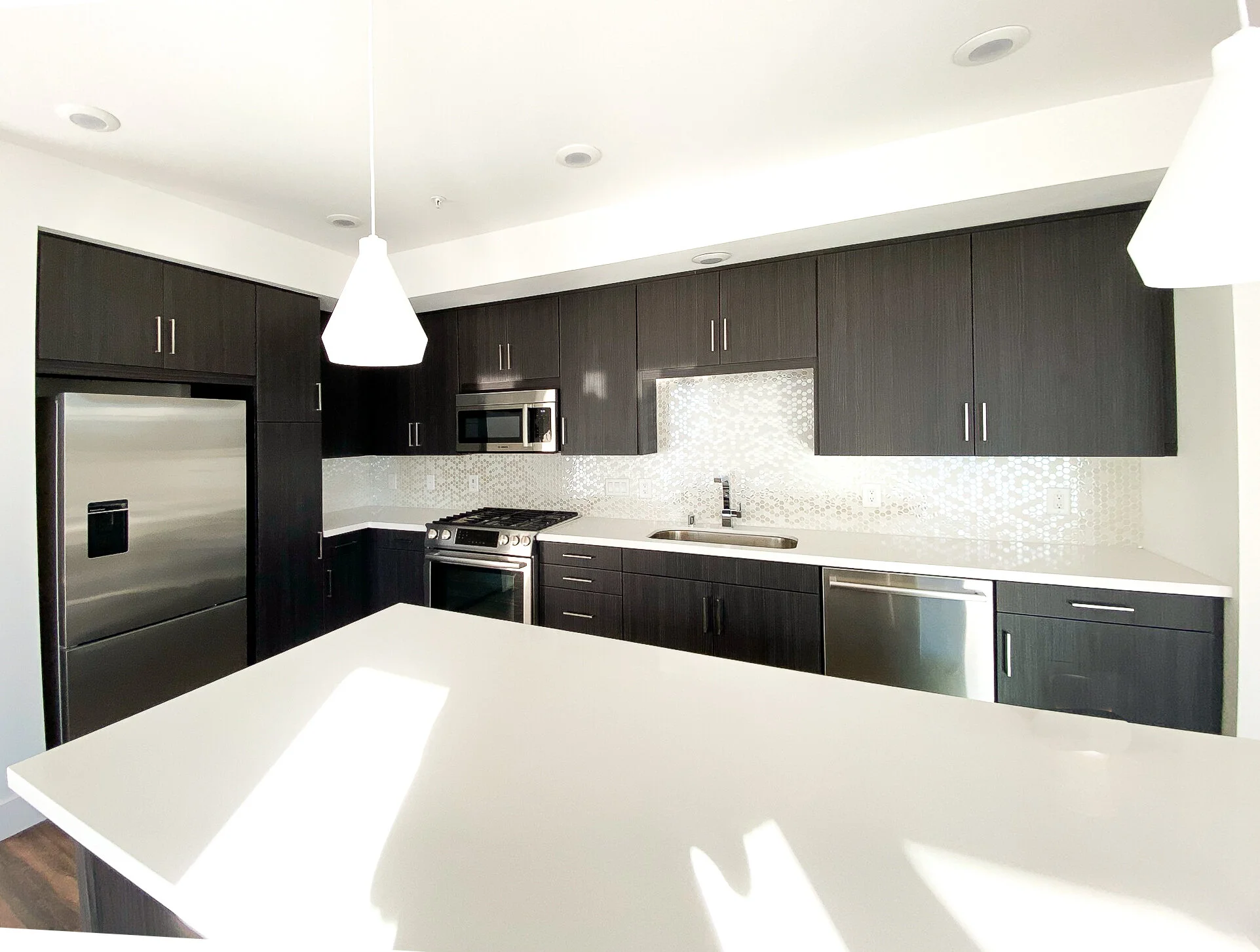 Modern kitchen with dark wood cabinets, stainless steel appliances, and white countertops, illuminated by hanging pendant lights.
