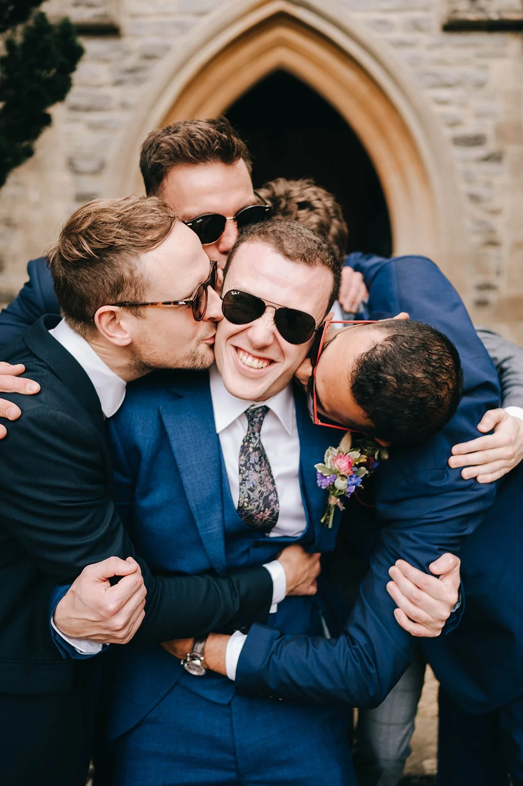 Groomsmen hugging and kissing the groom on his wedding day at Highnam Church in Gloucester