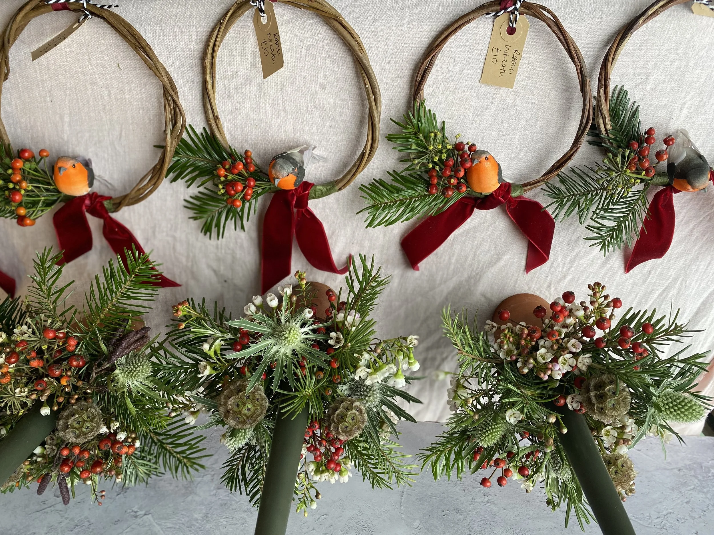 Decorative Christmas wreaths hanging on a wall, adorned with pine, holly berries, and red velvet ribbons, alongside bouquets of greenery with various flowers and berries.