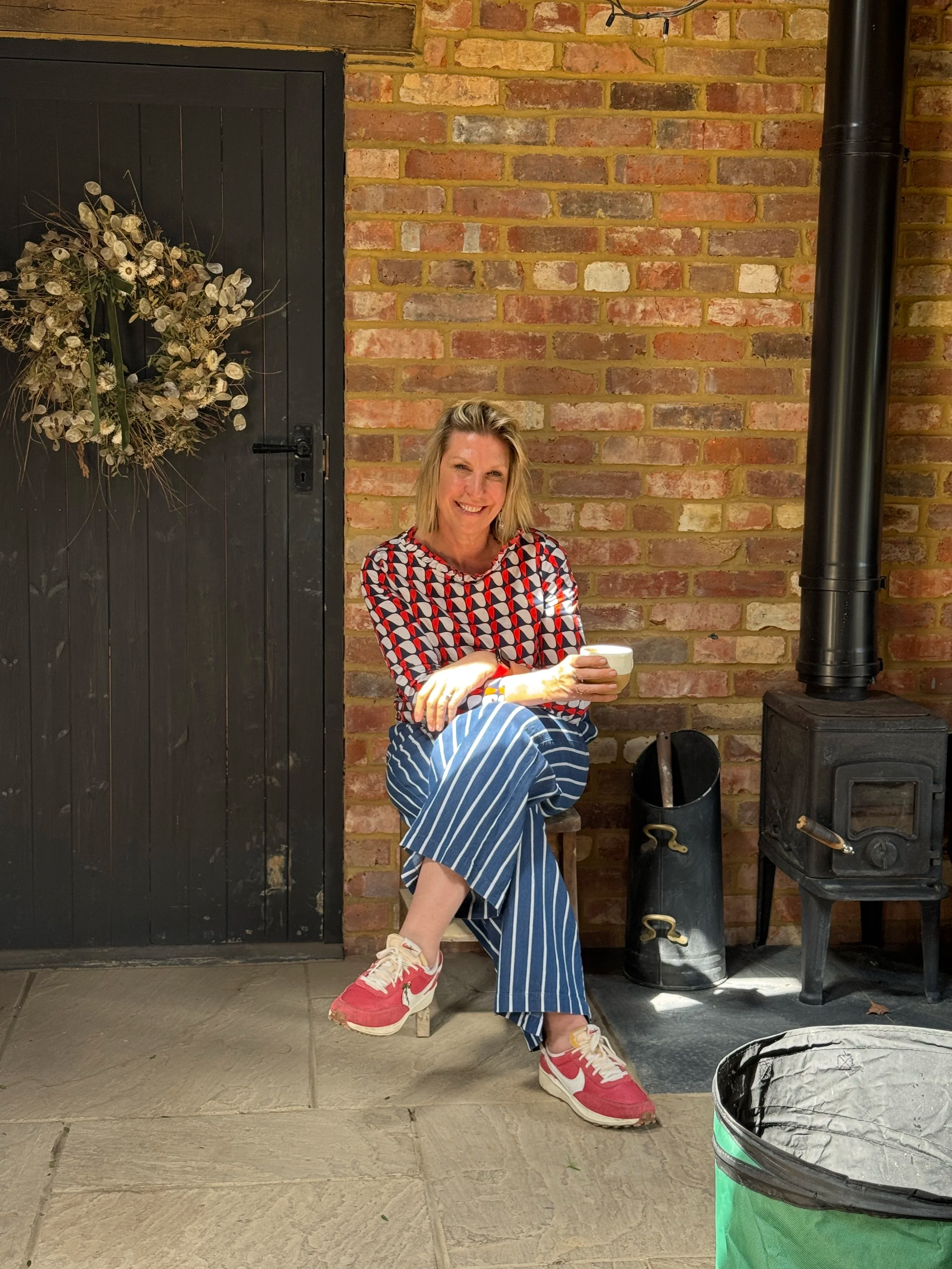 A smiling woman sitting on a small stool in front of a brick wall, holding a white mug. She is wearing a patterned top, striped pants, and pink sneakers. There is a wood stove and a black container beside her, with a green and black trash can in the foreground and a black door with a decorative wreath on it to her left.