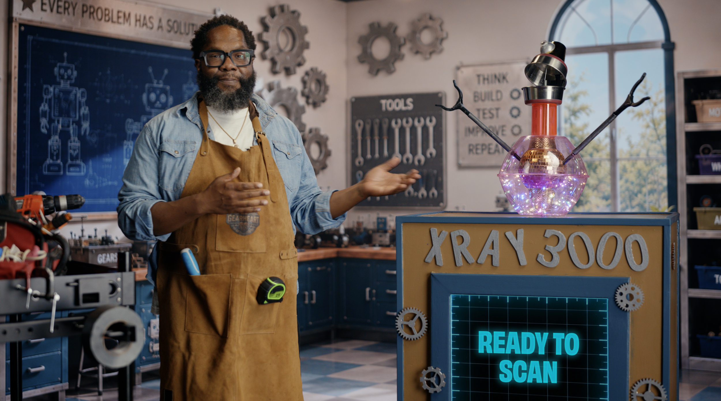 Man in denim shirt and apron explaining a science experiment in a workshop with tools and gears, next to a colorful X-ray machine labeled 'XRay 300' and a sign that says 'READY TO SCAN'.