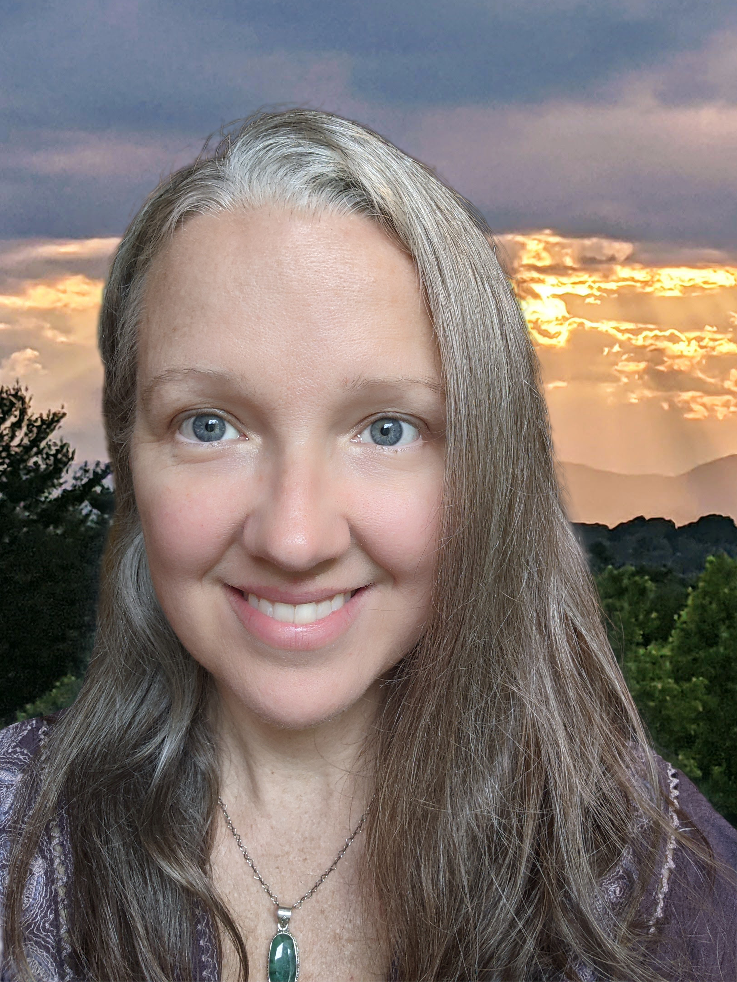A smiling woman with long brown hair and blue eyes, wearing a necklace with a green pendant, against a backdrop of a sunset sky with trees and mountains in the distance.