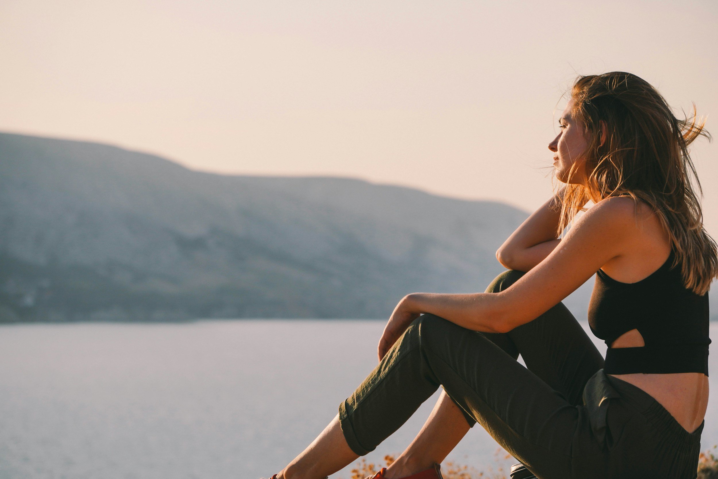 A woman with long hair sits on a rock, relaxing and enjoying the sunset near a body of water with mountains in the background.