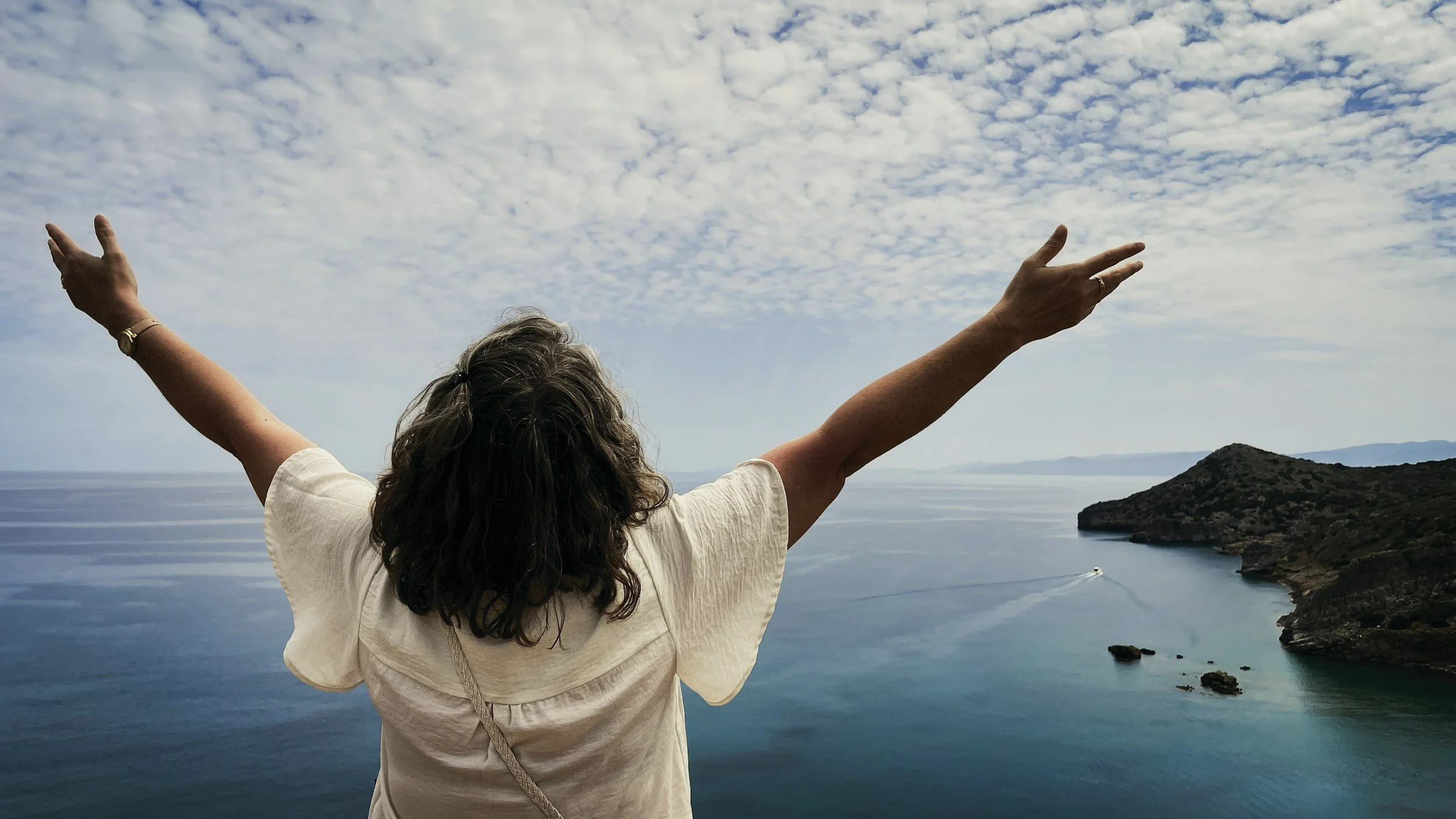 A woman with curly hair and a beige shirt, standing with arms outstretched, overlooking a body of water with a small island and a boat in the distance, under a partly cloudy sky.
