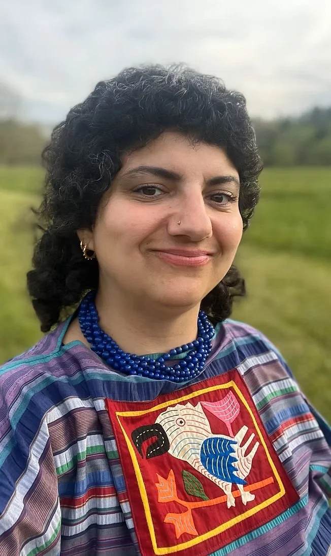 A woman with short curly black hair, wearing a nose ring, earrings, blue beaded necklace, and colorful striped top with an embroidered bird design on the front, standing outdoors in a grassy field under a cloudy sky.