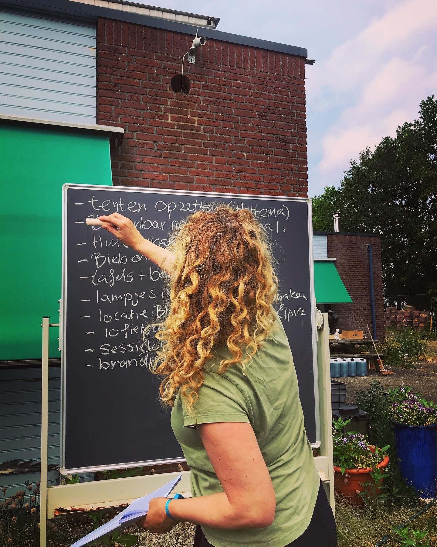 Een vrouw met krullend blond haar schrijft op een schoolbord met witte krijt buiten, met een groen en een brick gevel op de achtergrond en planten op de voorgrond.