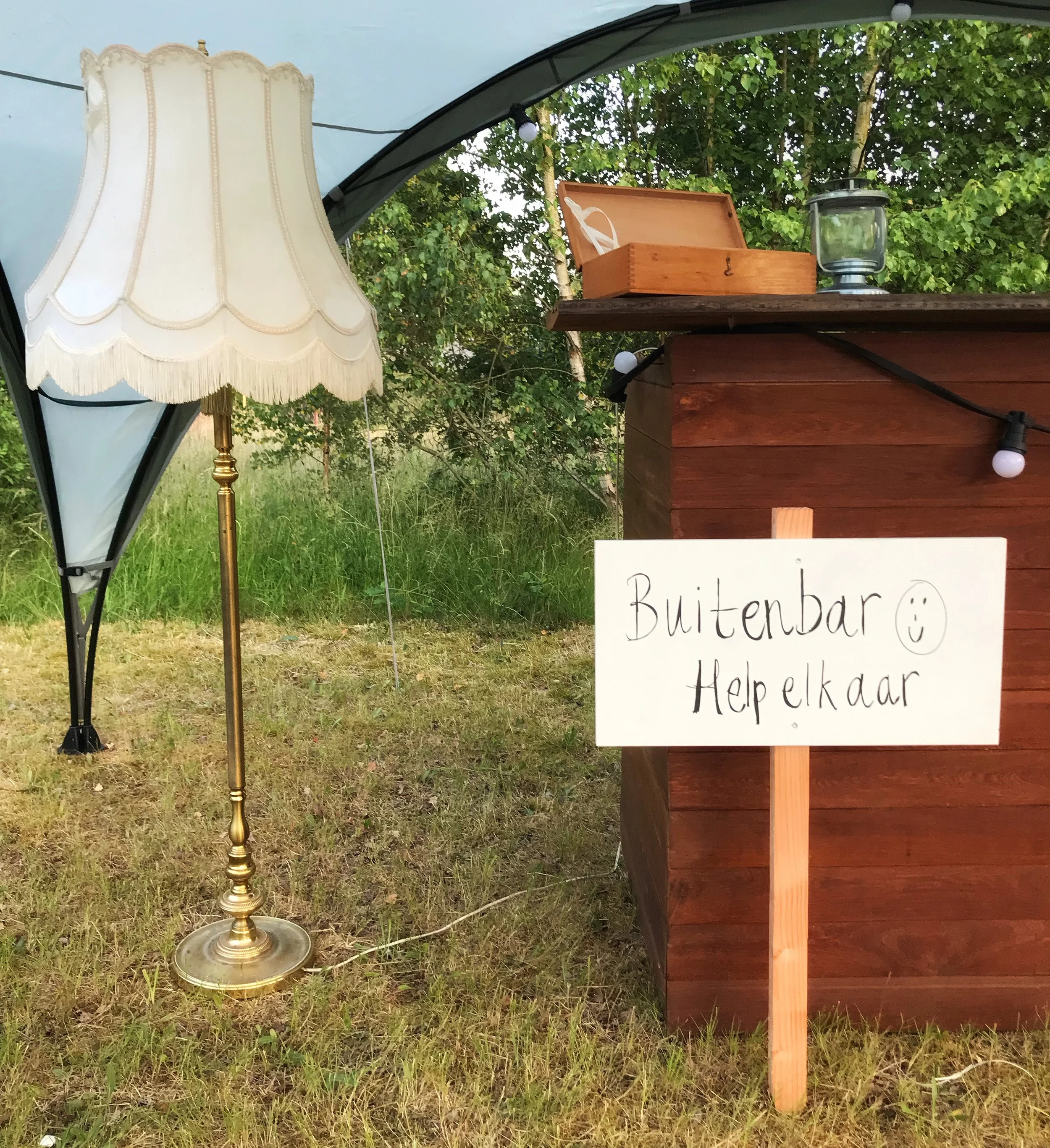 Een buitenbar met een houten uitstraling, een lamp, een houten kist en een bord met de tekst 'Buitenbar. Help jezelf'.