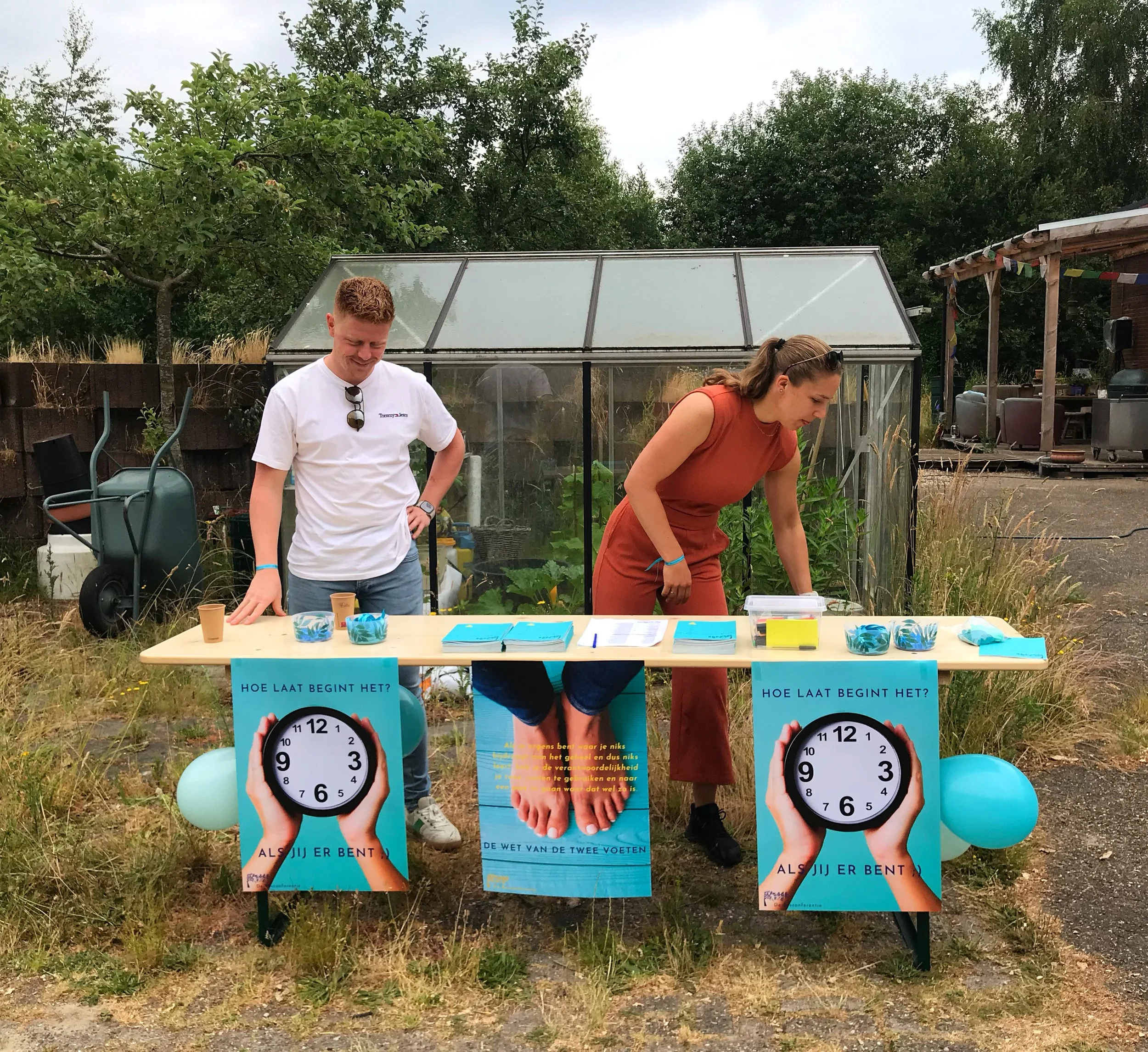 Twee mensen staan achter een tafel met posters en flyers op een grasveld, met een kas en bomen op de achtergrond. De posters tonen een klok en tekst in het Nederlands.