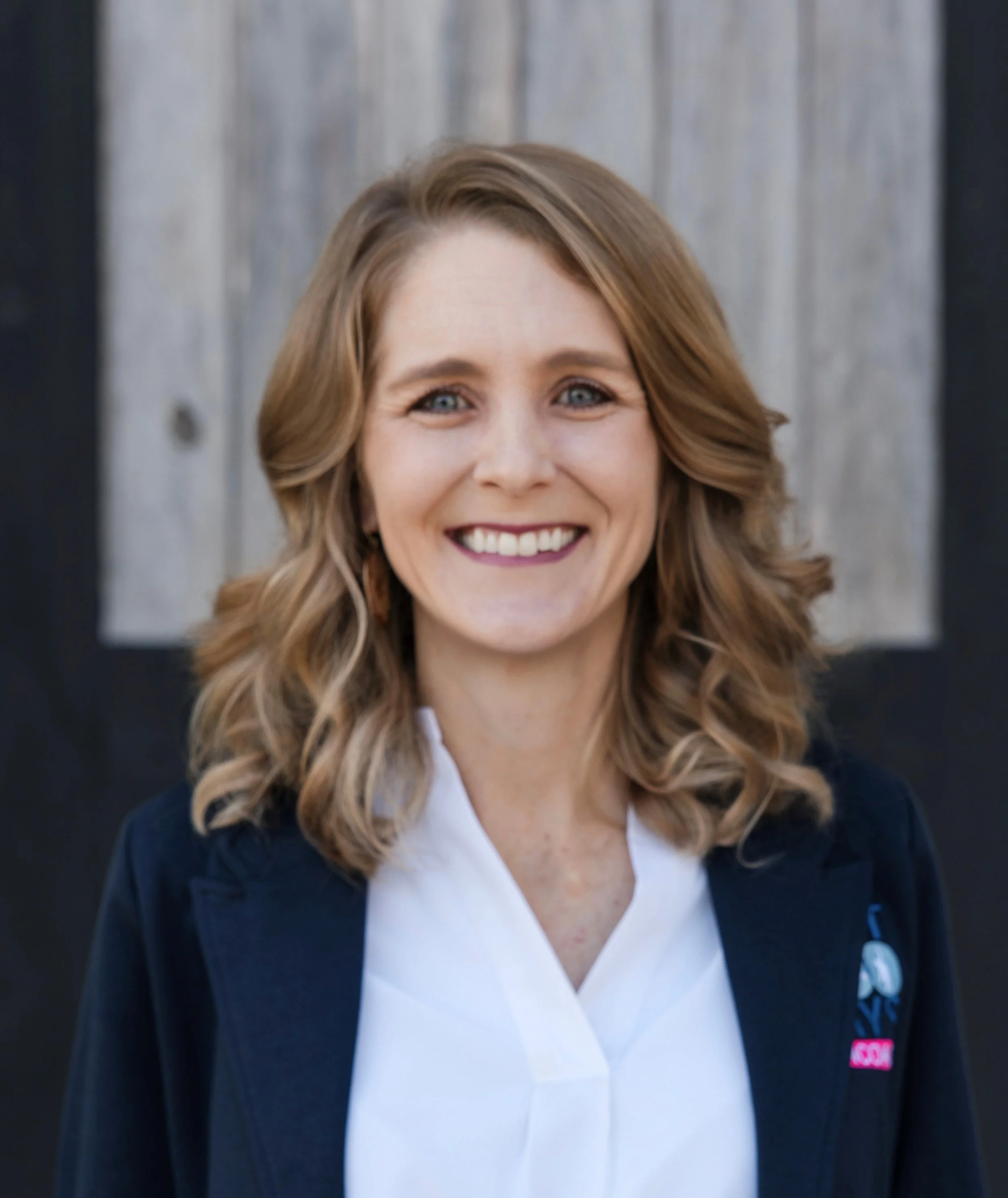 A smiling woman with long wavy light brown hair, wearing a navy blue blazer over a pink blouse with a matching pink scarf, and a silver necklace with a blue pendant, standing outdoors with blurred green trees in the background.