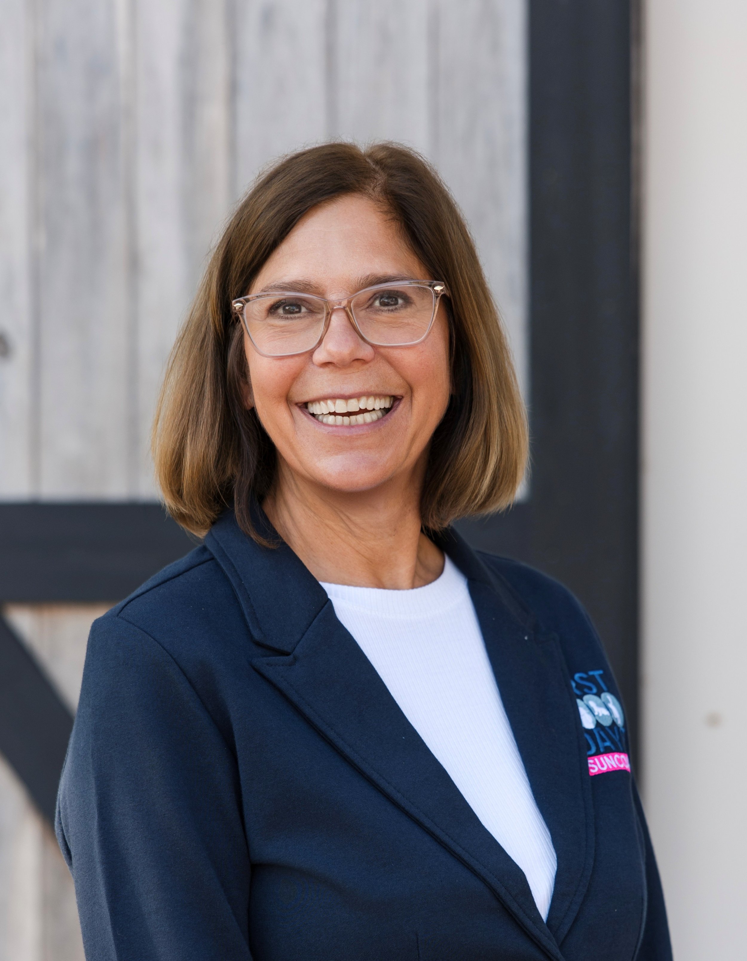 A woman with shoulder-length brown hair, glasses, and a bright smile, wearing a dark blazer and a pink top, standing outdoors with trees and a cloudy sky in the background.