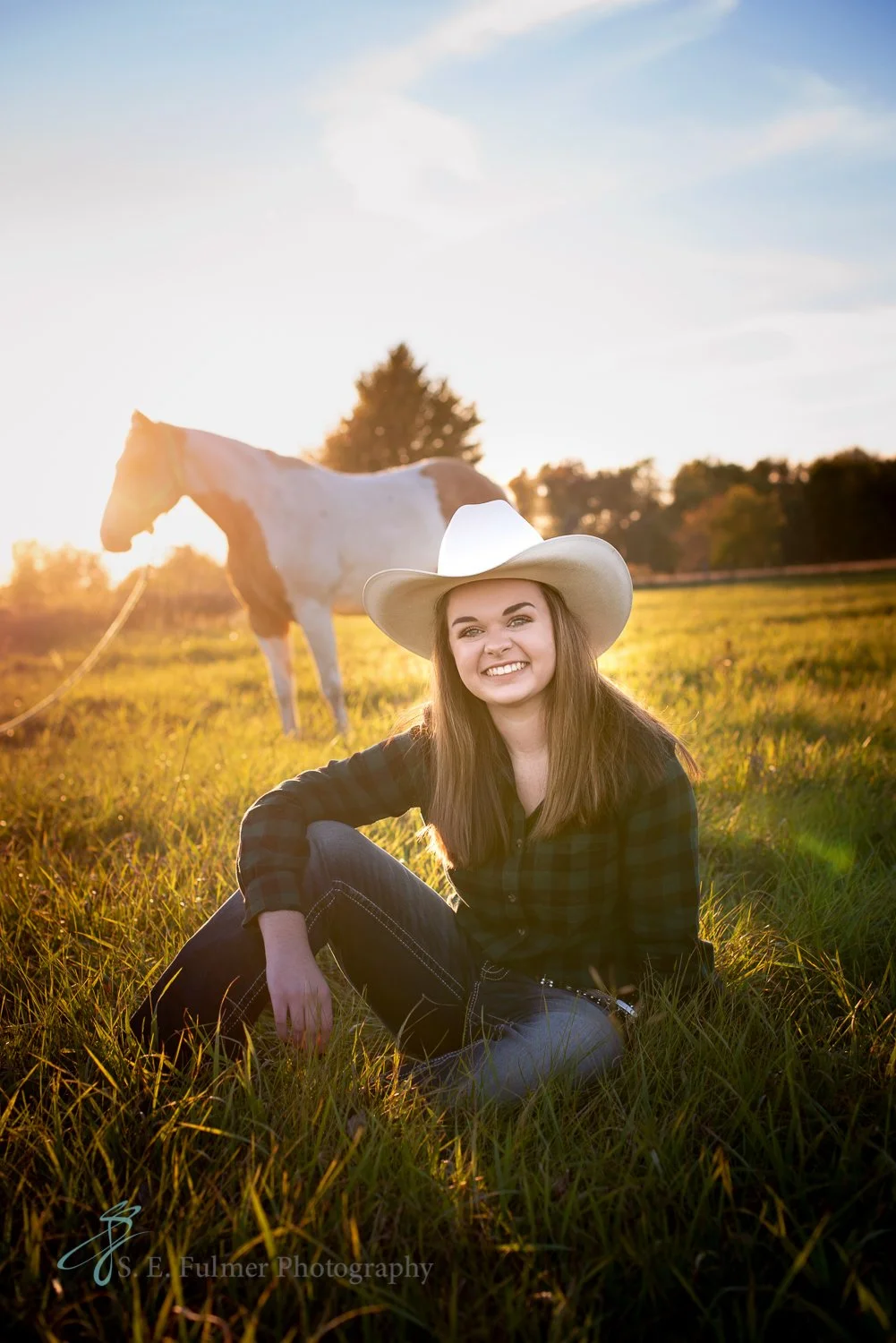 High school senior, Goodrich, MI.  Young woman wearing a cowboy hat sitting in a grassy field with a horse in the background at sunset. North Branch, MI.