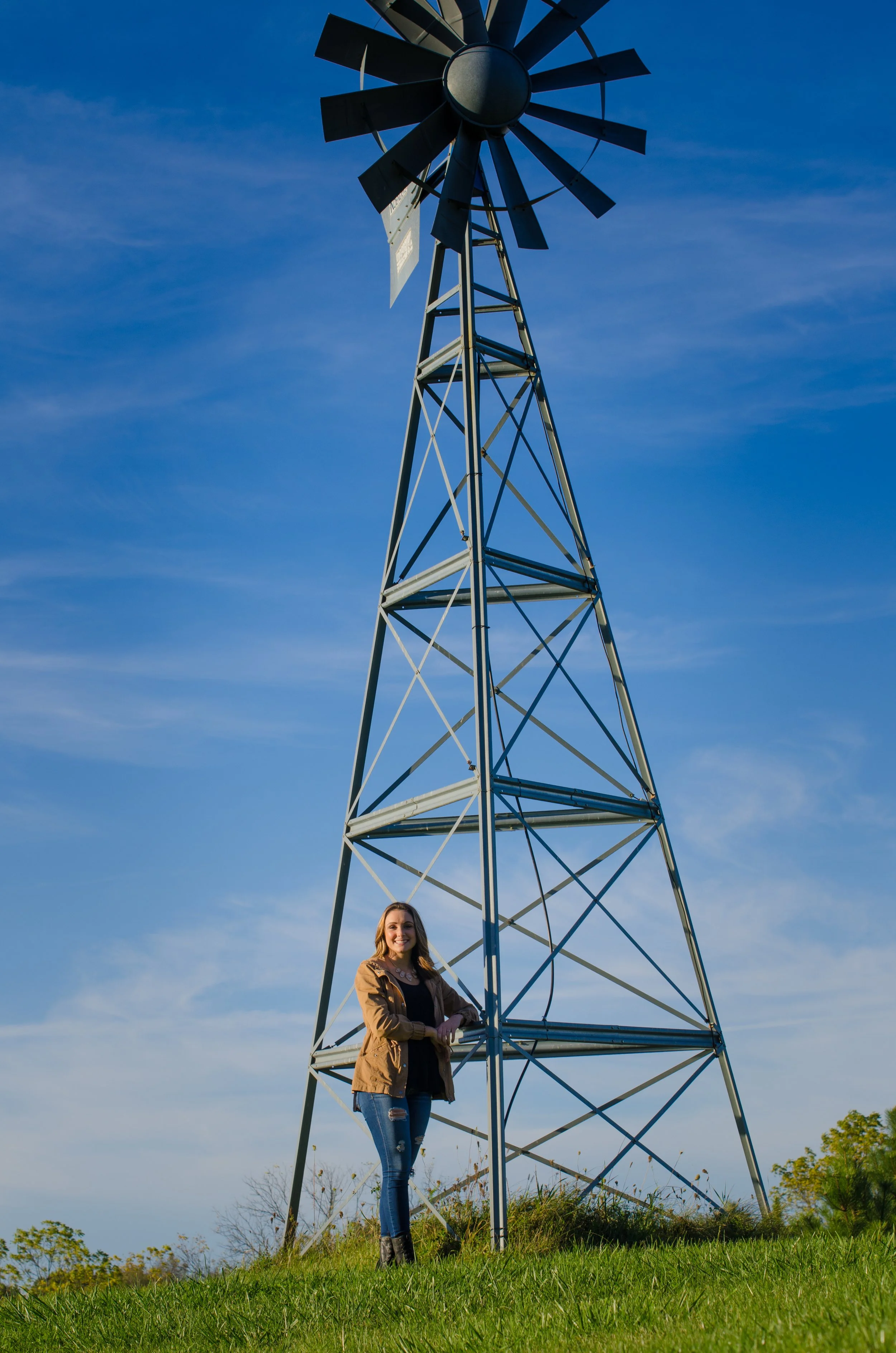 High school senior, Goodrich, MI. A woman standing in front of a windmill tower on a grassy field under a blue sky.