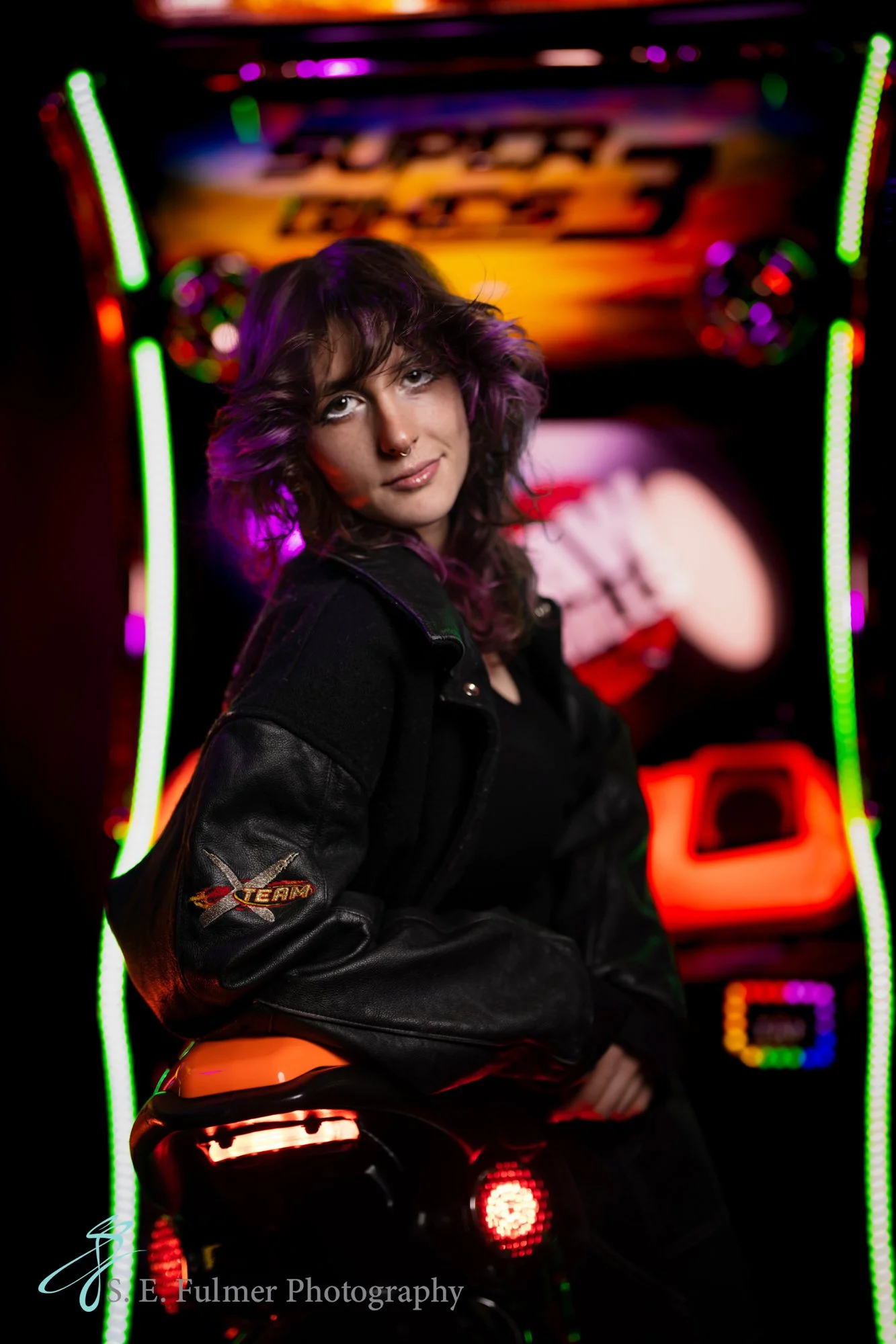 High school senior, Goodrich, MI. Girl in front of arcade game wearing a leather jacket with colorful lights.