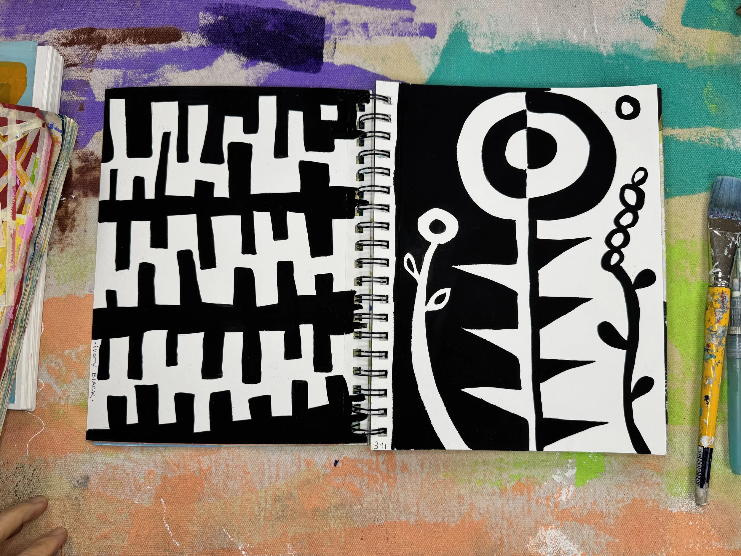 Open sketchbook displaying two abstract black and white designs, one on each page, with markers and art supplies visible on a colorful, textured table.
