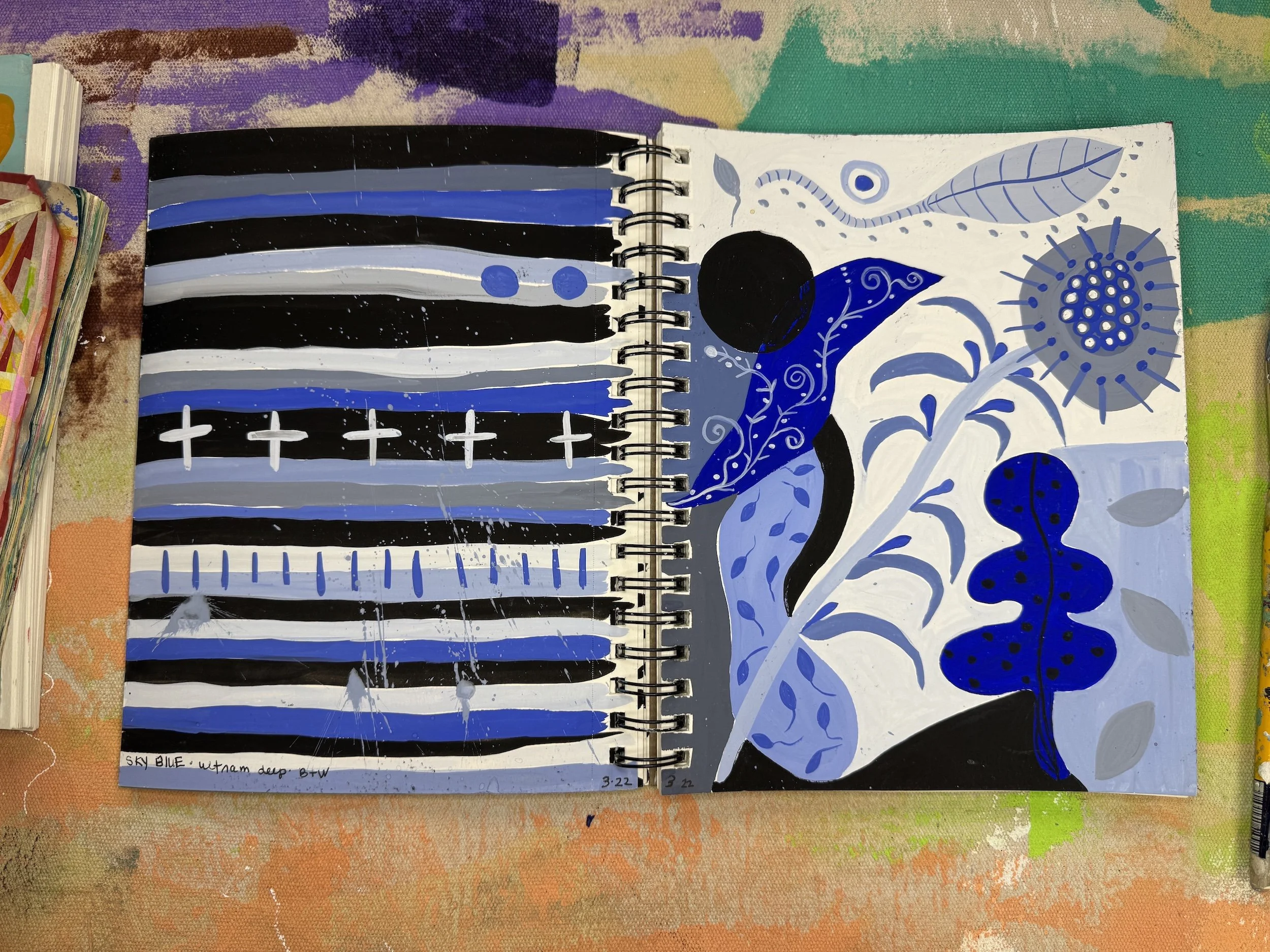 Open spiral notebook with abstract blue, black, white, and gray artistic designs on two pages. The left page features horizontal stripes, some with crosses and yellow splatters. The right page has stylized plants, leaves, and circular shapes with swi