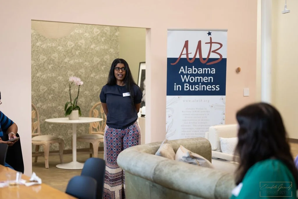 A woman standing in front of a banner that reads "Alabama Women in Business" during a meeting or event, with other women seated and listening.