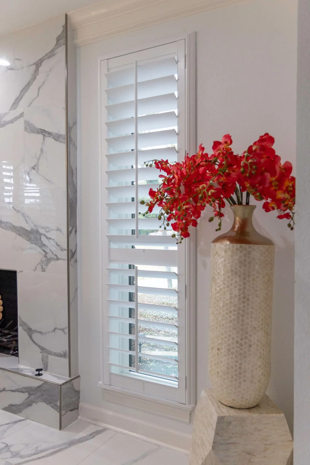 Tall window in Dallas urban home entry with plantation shutters in bright white. Large vase with colorful flowers sits next to the window.