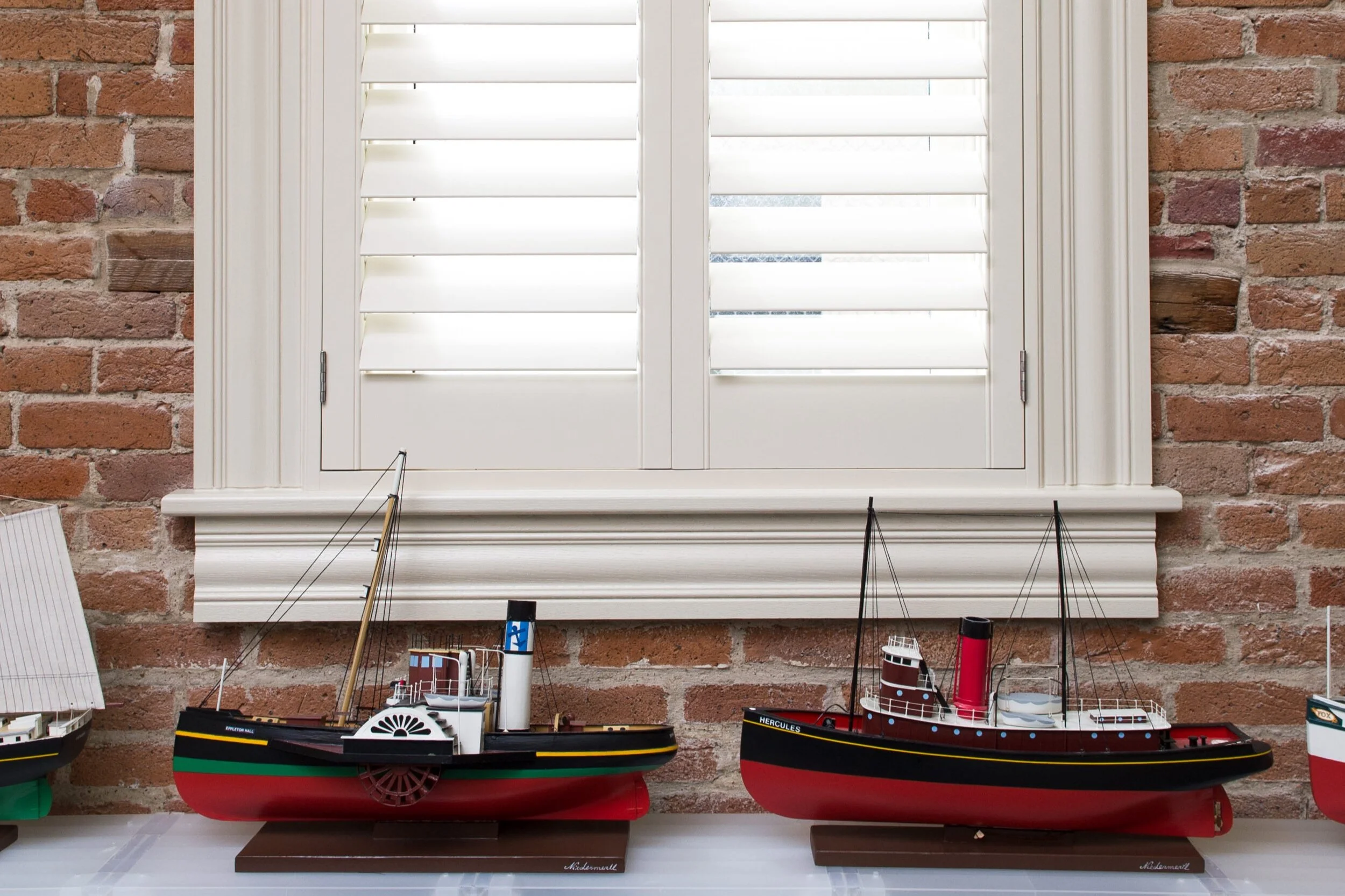A window with white shutters with model ships displayed below.