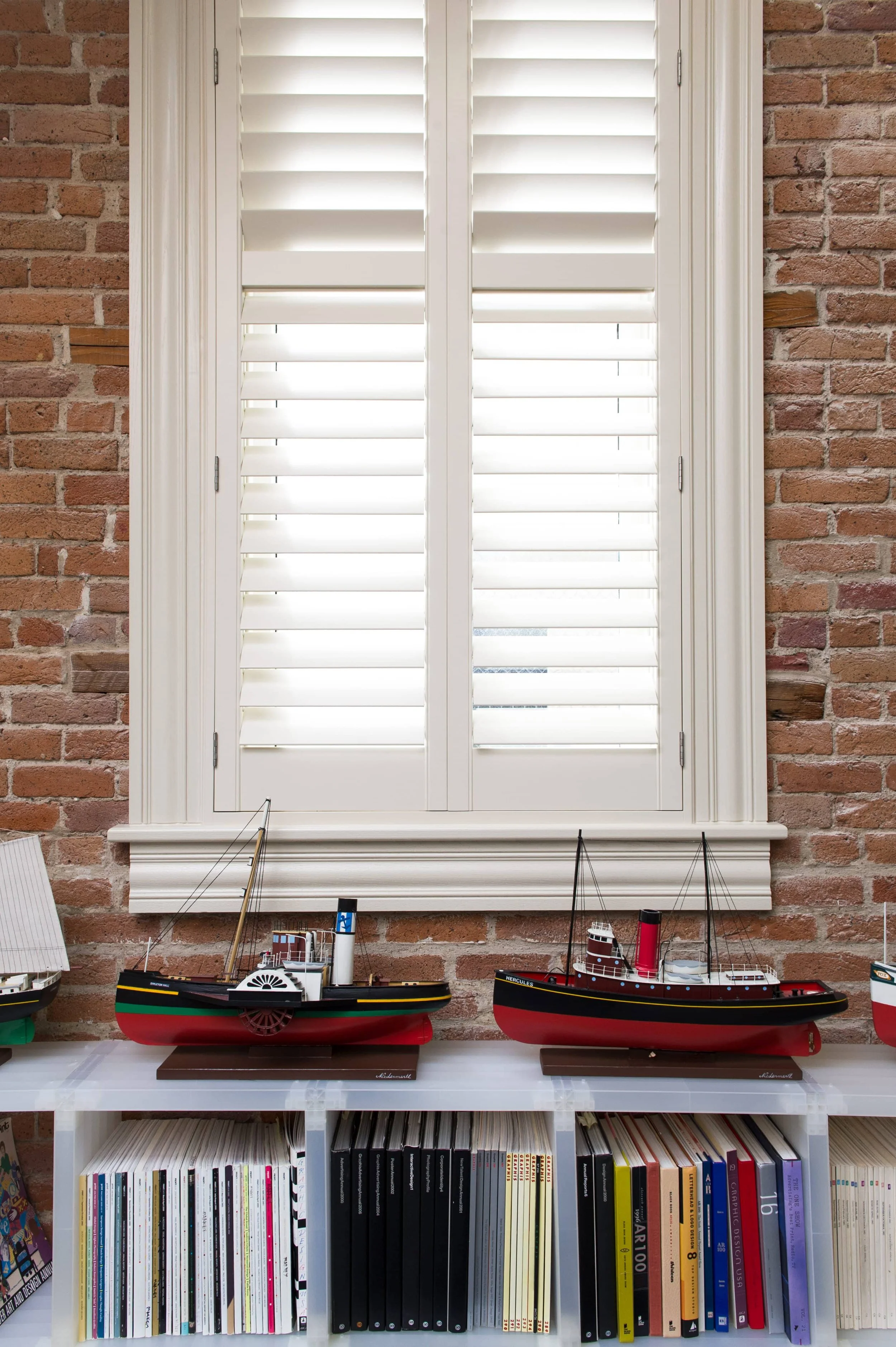 White custom shutters framed by exposed brick above a display of model fireboats in a St. Louis firehouse.