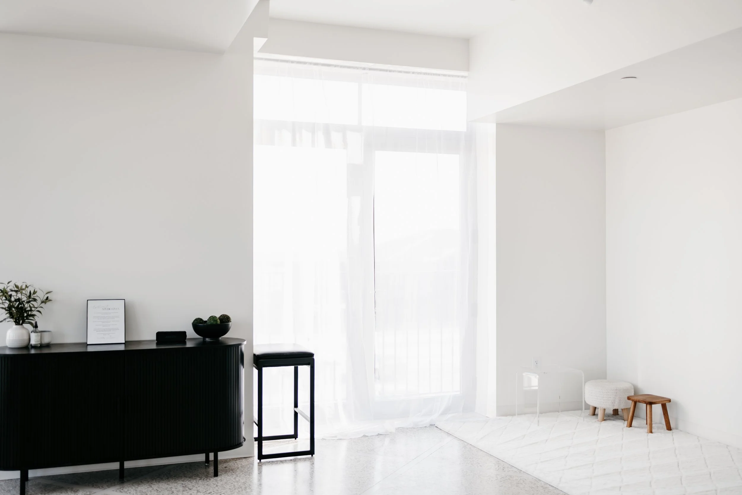 Bright, minimalist living space with white walls, sheer curtains, a black sideboard with decorative items, and small seating stools on a white rug.