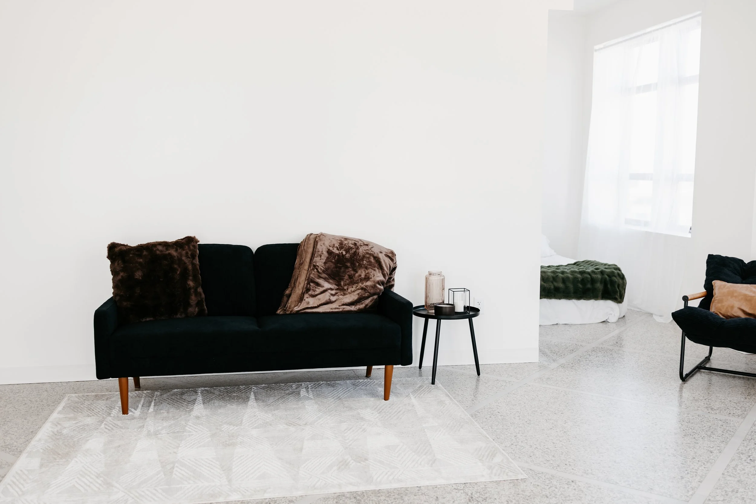 Living room with a black sofa, a side table, and throw pillows, adjacent to a bedroom visible through an open space, with a bed covered in a green blanket and a black lounge chair with throw pillows.