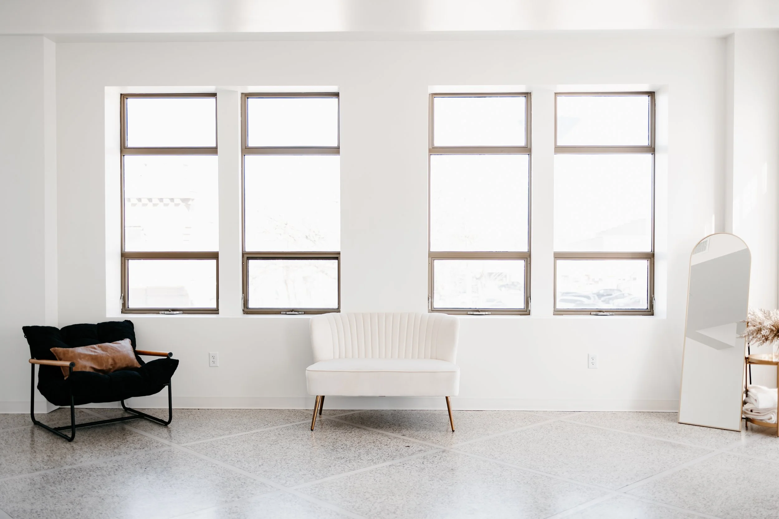 Minimalist interior with three large windows, a white cushioned bench, a black armchair with a brown pillow, and a full-length mirror on a stand near a small side table with dried flowers.