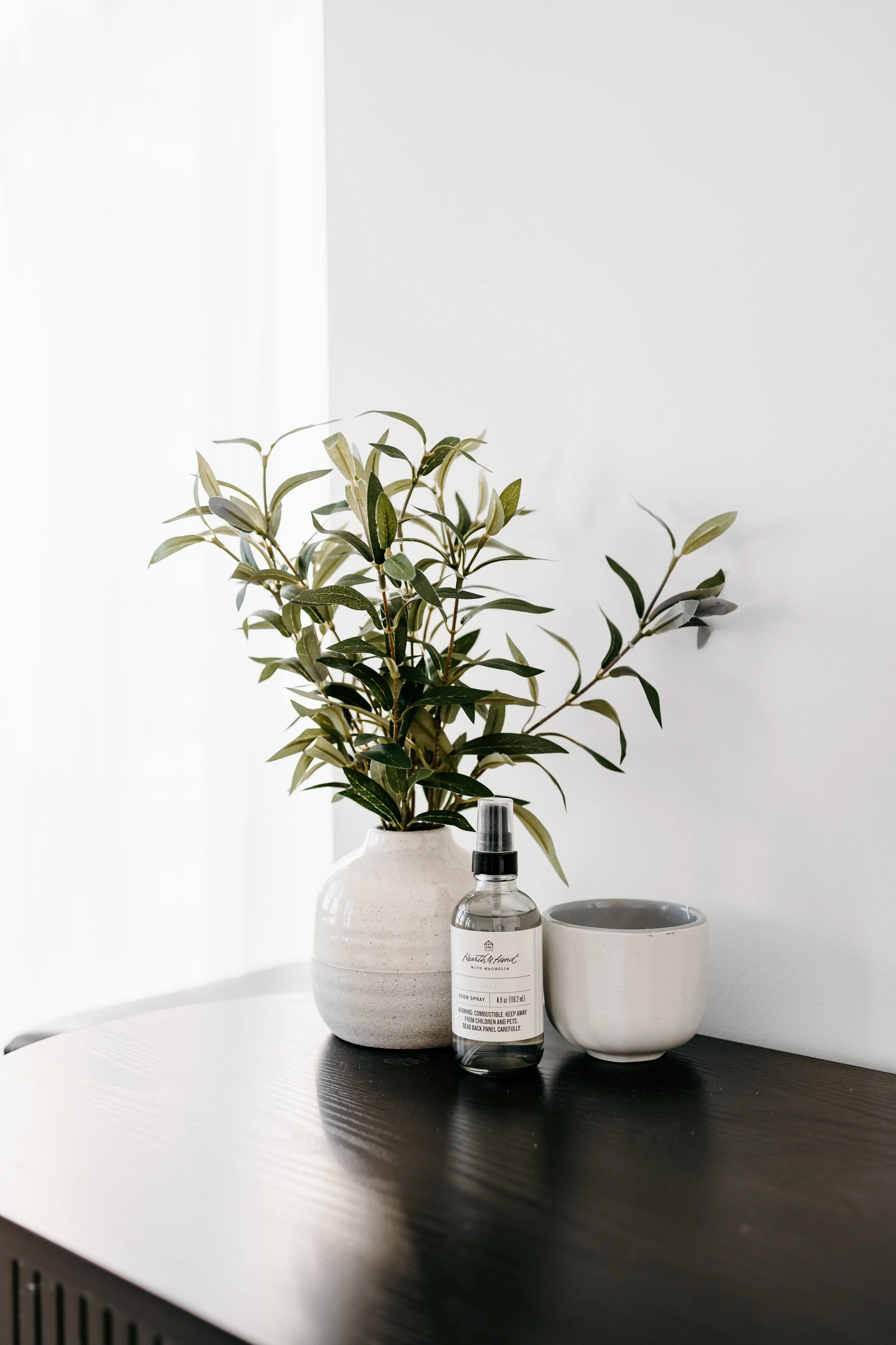 A white vase with green leafy plant, a spray bottle of room spray, and an empty white ceramic bowl on a dark wooden table against a white wall.