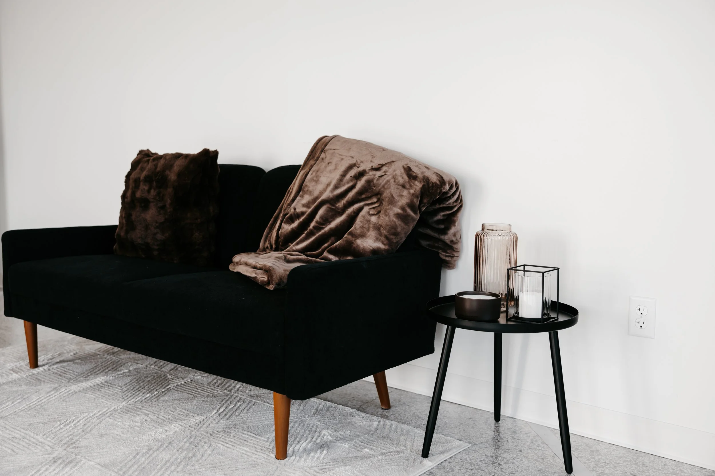 A black sofa with two brown throw pillows and a brown blanket, placed against a white wall. Beside the sofa is a small black side table with a tall glass vase, a candle holder with a white candle, and a small brown bowl. There is an electrical outlet