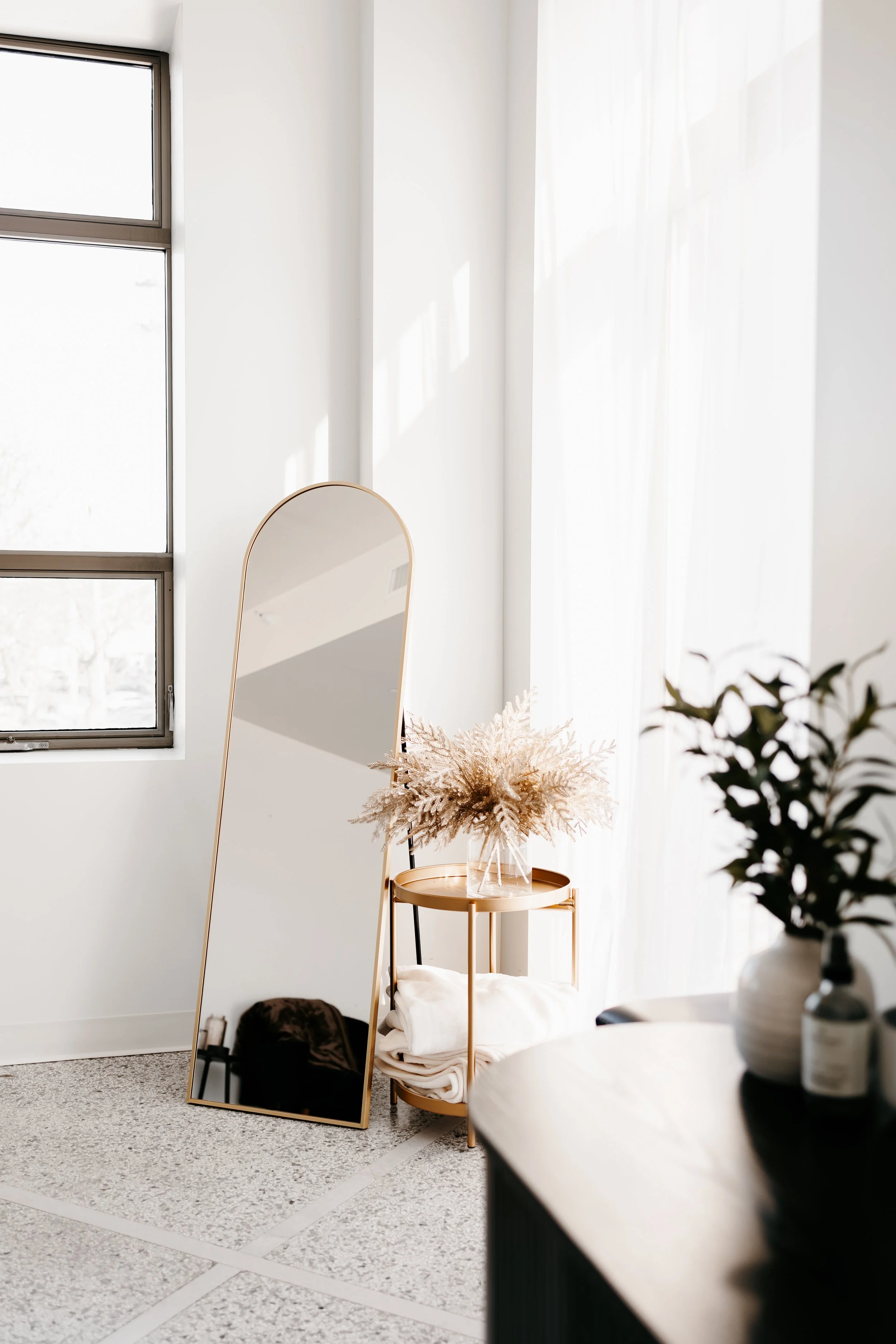 A minimalist room corner with a large standing mirror, a small side table holding a glass vase with pampas grass, a folded blanket, a potted plant, and sheer white curtains illuminated by natural sunlight.