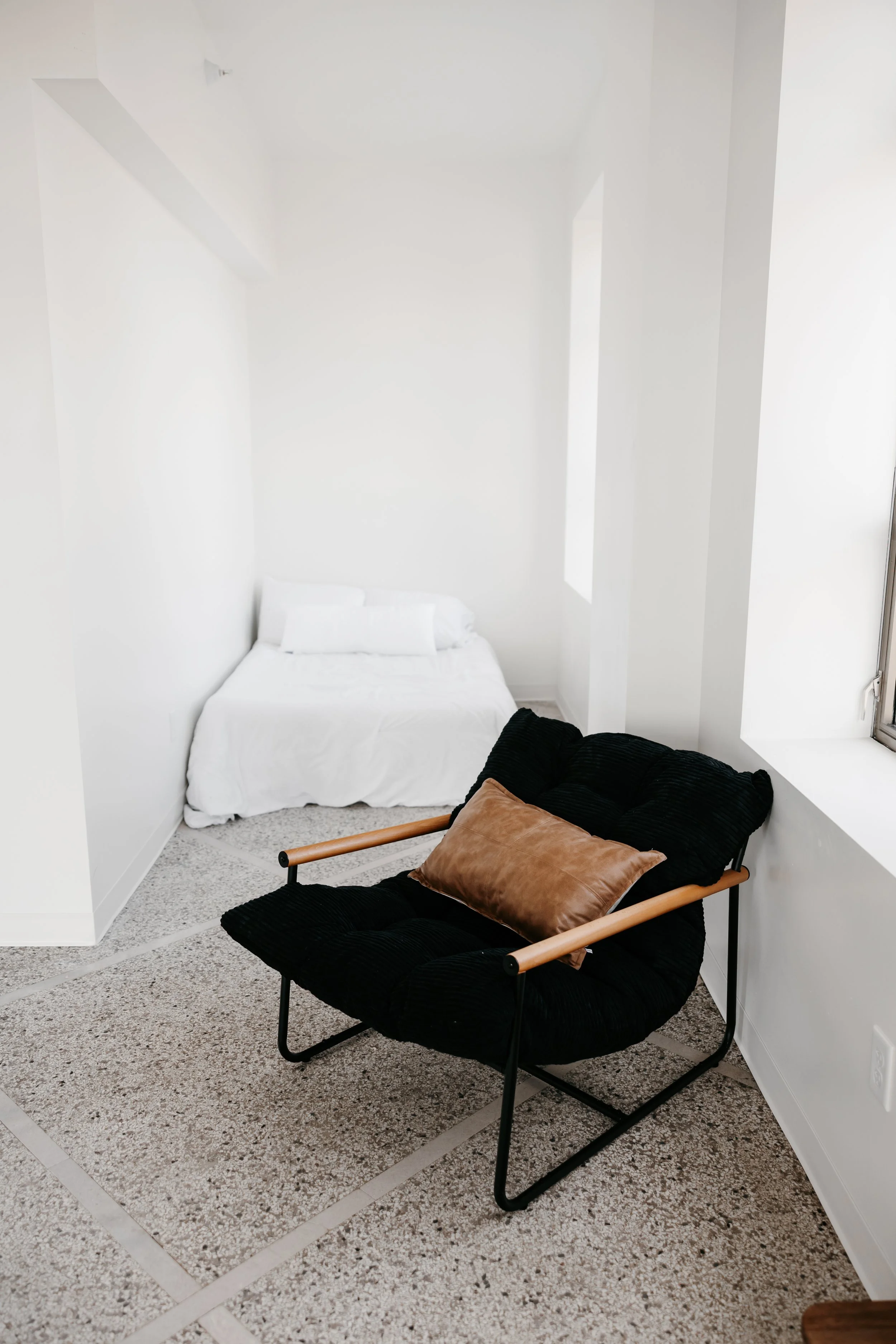 A small room with white walls and a bed covered in white linens in the back corner. A black cushioned armchair with wooden armrests and a brown throw pillow is in the foreground near a window.