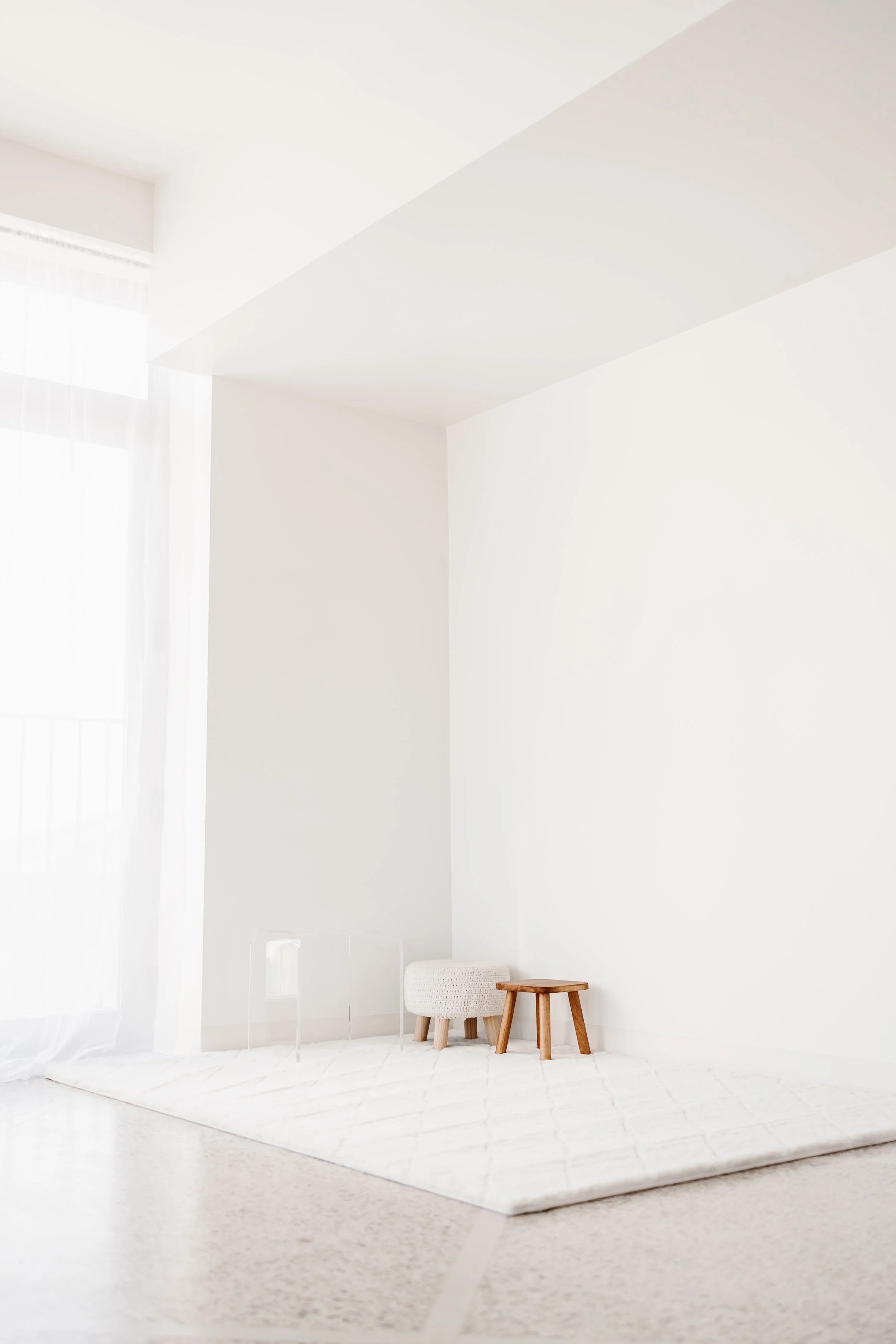 Minimalist white room with a white rug, small furniture including a knitted footstool, a wooden stool, and a transparent side table, with a large window and sheer curtains.
