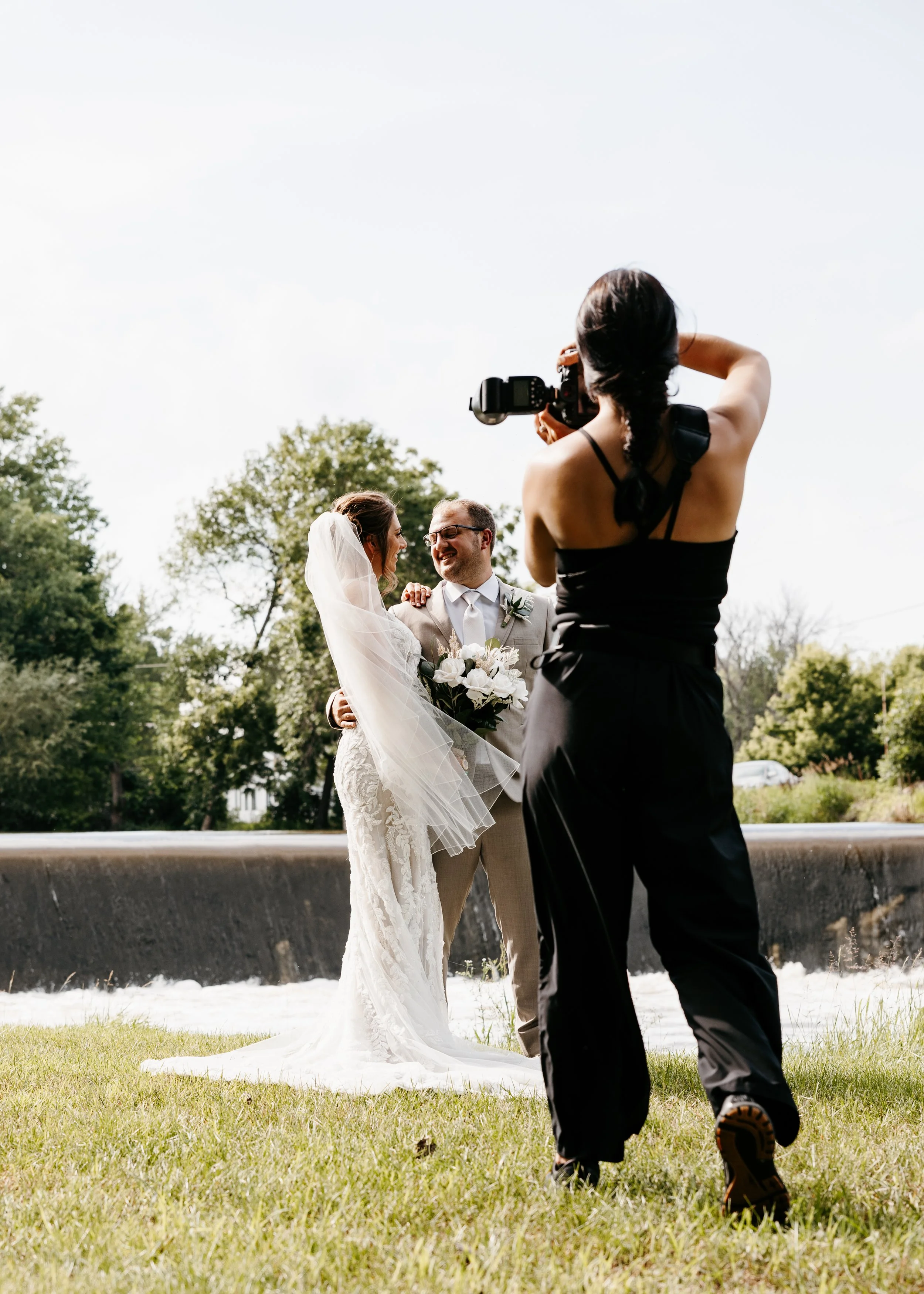A woman in a black dress takes a photo of a bride and groom during their wedding outdoors. The bride is wearing a white wedding dress with a veil, and the groom is in a light-colored suit holding a bouquet of flowers. They are smiling at each other, standing near a body of water, with trees in the background.