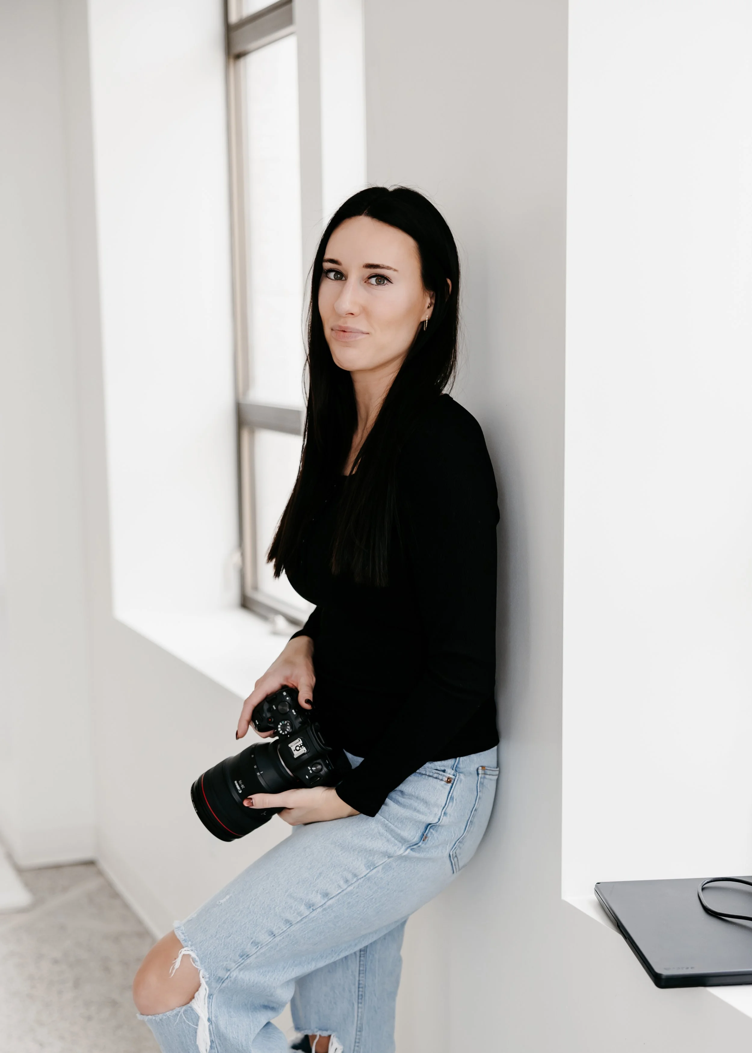 A woman with long black hair, wearing a black long-sleeve shirt and light blue ripped jeans, is standing in a bright room, holding a professional camera with a large lens, and leaning against a white wall near a window.